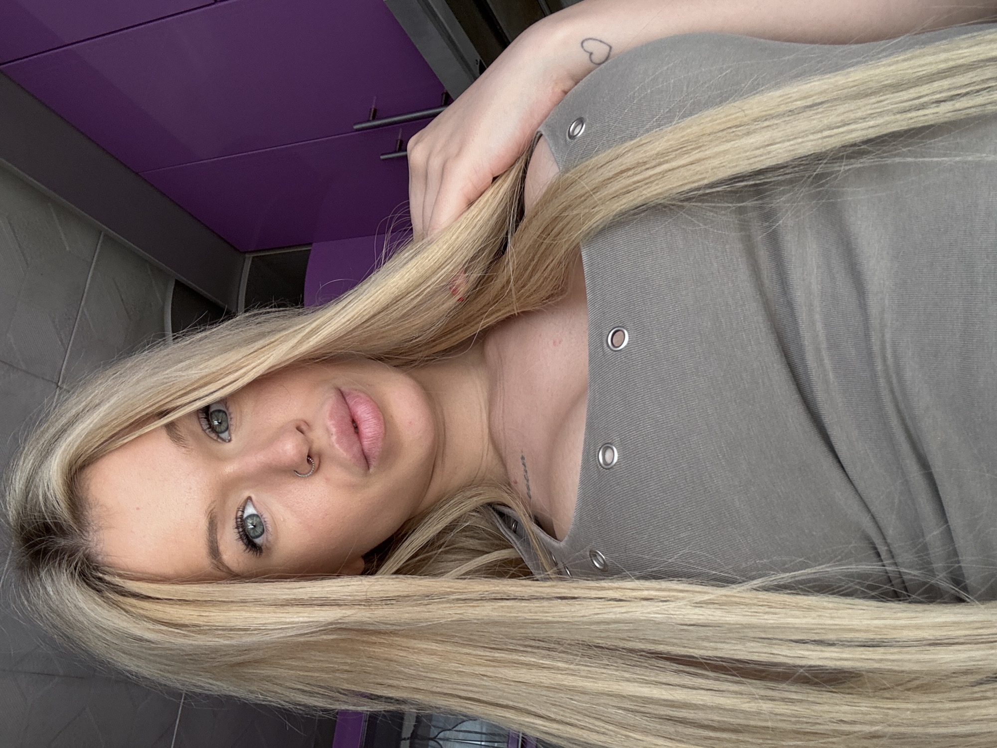 The media file is a close-up, portrait-style photograph of a young Caucasian woman with long, blonde hair. She is wearing a gray, scoop-neck t-shirt with visible eyelet detailing on the shoulders. Her hair is styled long and straight, falling around her face and shoulders. She has blue eyes, visible eyelashes and eyebrows, and full lips. A nose ring is visible on her left nostril. A small, outlined heart tattoo is on her left forearm, partially obscured by her hair and arm. She is looking directly at the camera.
The background suggests an indoor setting, possibly a kitchen or bathroom, with elements like purple cabinets and grey tiled walls visible. The lighting appears to be natural, coming from the front, and the time of day is not definitively indicated. There is no discernible weather. The overall mood is casual and relaxed. There is no visible text in the image.