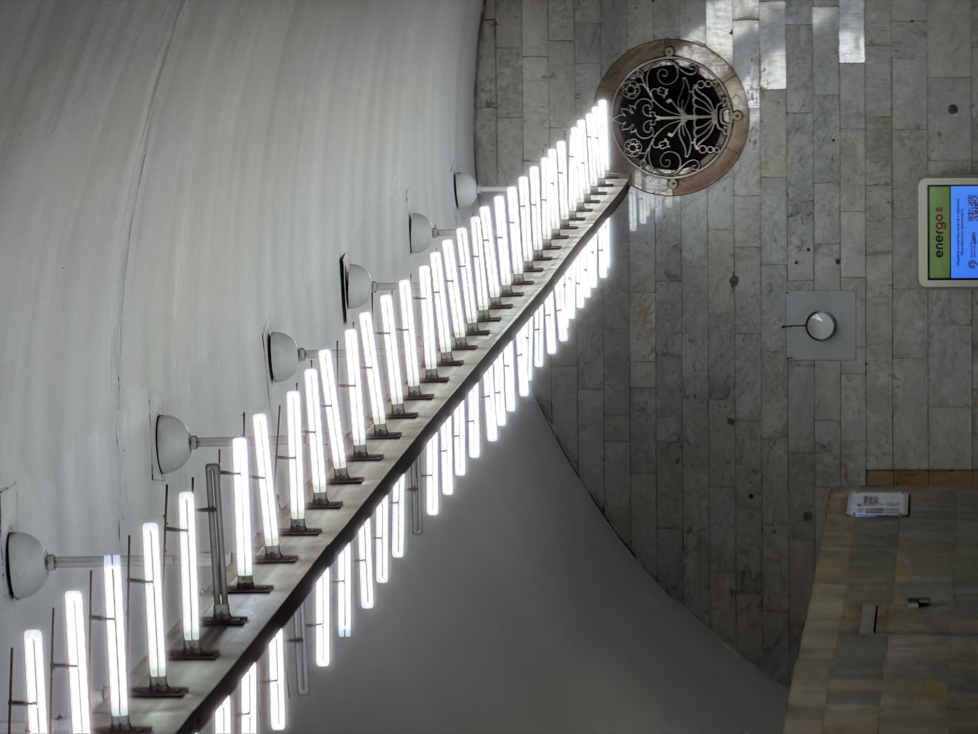 This is an interior shot of the Mayakovskaya metro station in Moscow, Russia. The view is upward, featuring a row of illuminated cylindrical lights attached to a horizontal beam extending diagonally across the image. The lights are mounted on metal brackets affixed to a curved white wall on the left side of the image.
On the right side of the image, a wall constructed of vertical rectangular tiles is visible. At the top, there is a decorative circular ventilation grill. Farther down the wall, a digital display shows information in a foreign language (likely Russian). A lighter colored, angular structure intersects the right wall in the lower portion of the image. The light source is internal from the tubes.