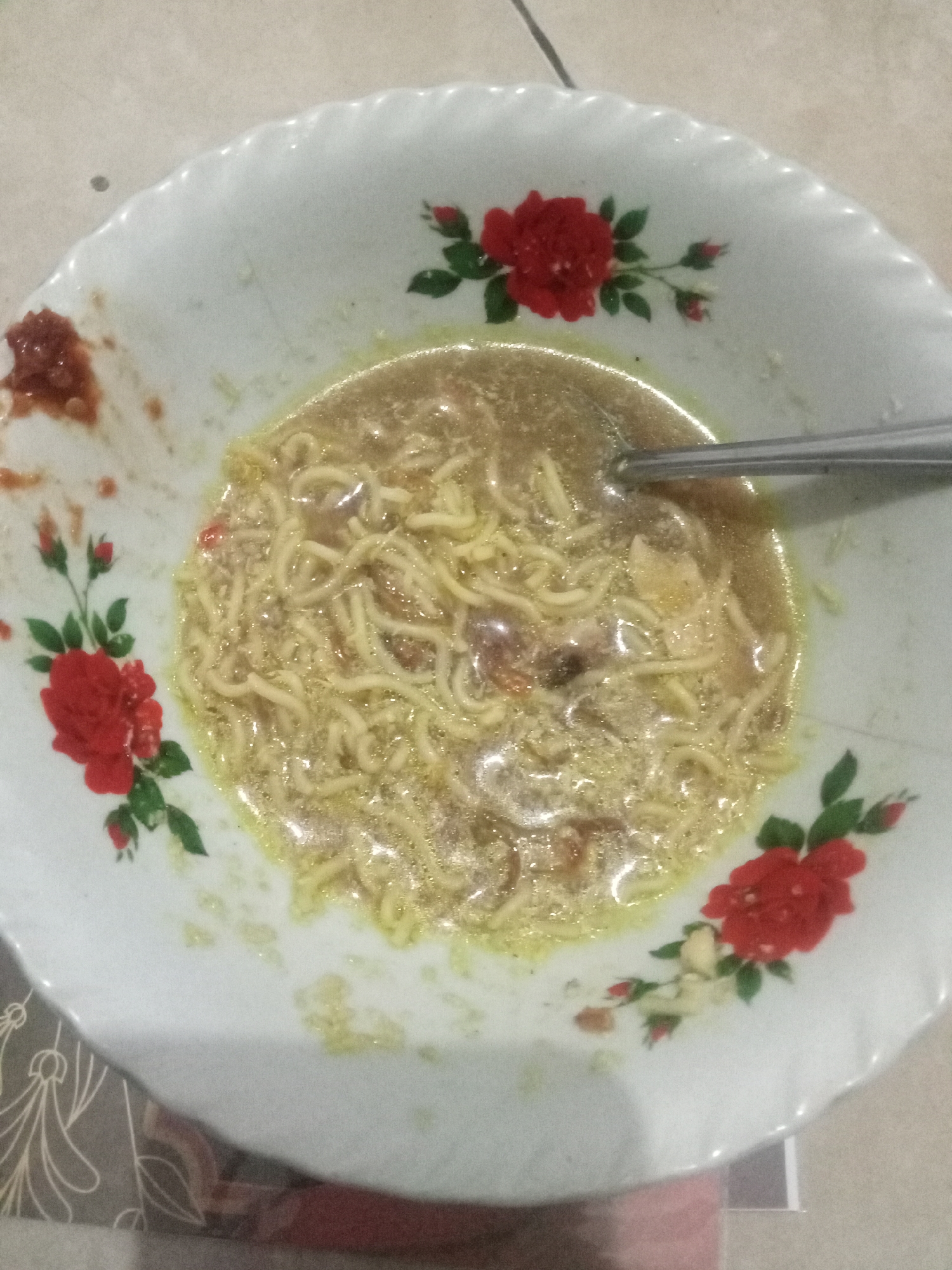 A top-down, close-up shot shows a bowl of instant noodles with broth. The bowl is white with a scalloped edge and decorated with red roses. A silver spoon is partially submerged in the noodles. To the left of the bowl, there is a small dollop of red chili paste. The background appears to be a tiled surface. The image was taken in the city of Malang, Indonesia.
