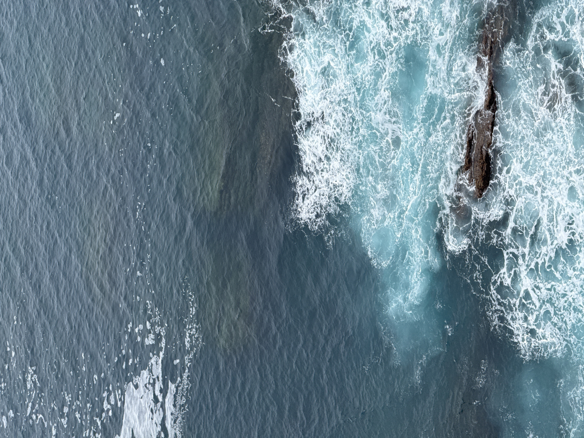 This image depicts a close-up, overhead view of the ocean waters near the town of Cudillero, Spain. The sea is a deep blue-green, with areas of lighter turquoise where waves are breaking. White foam and spray are visible as waves crash against dark, submerged rocks and the shoreline. The water's surface is rippled, indicating moderate movement. No people or structures are visible in this natural scene.