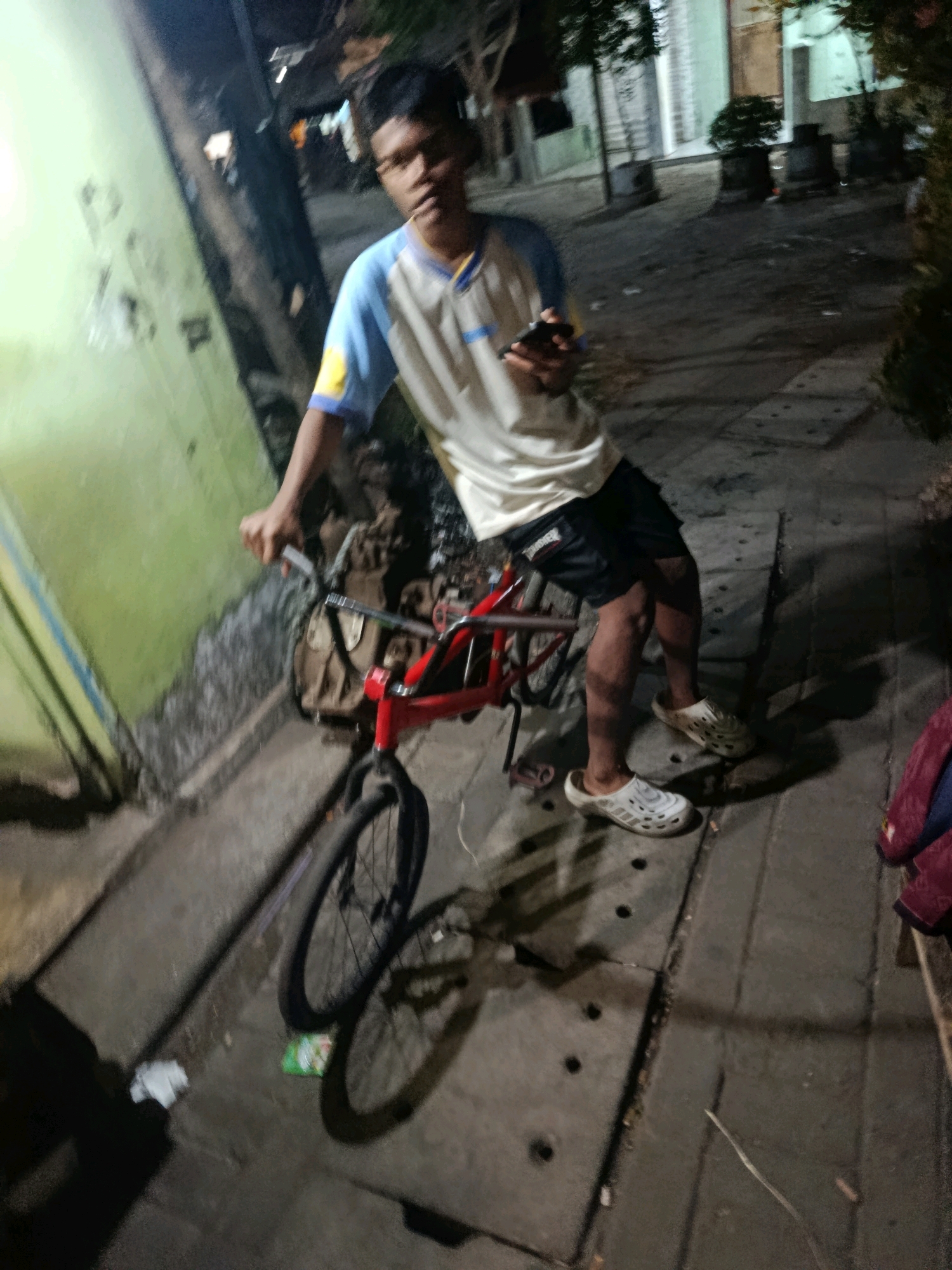 A young male, appearing to be of Indonesian descent, stands next to a red bicycle on a paved sidewalk at night. He is wearing a white and blue t-shirt, black shorts, and white slip-on shoes. The bicycle has a brown bag attached to its frame. The background shows buildings, trees, and street lighting, suggesting an urban environment in Jobokuto, Indonesia. The lighting is dim, typical of nighttime. The individual is holding a mobile phone in his right hand. The scene is captured from a slightly low angle, with motion blur indicating movement or a quick capture.