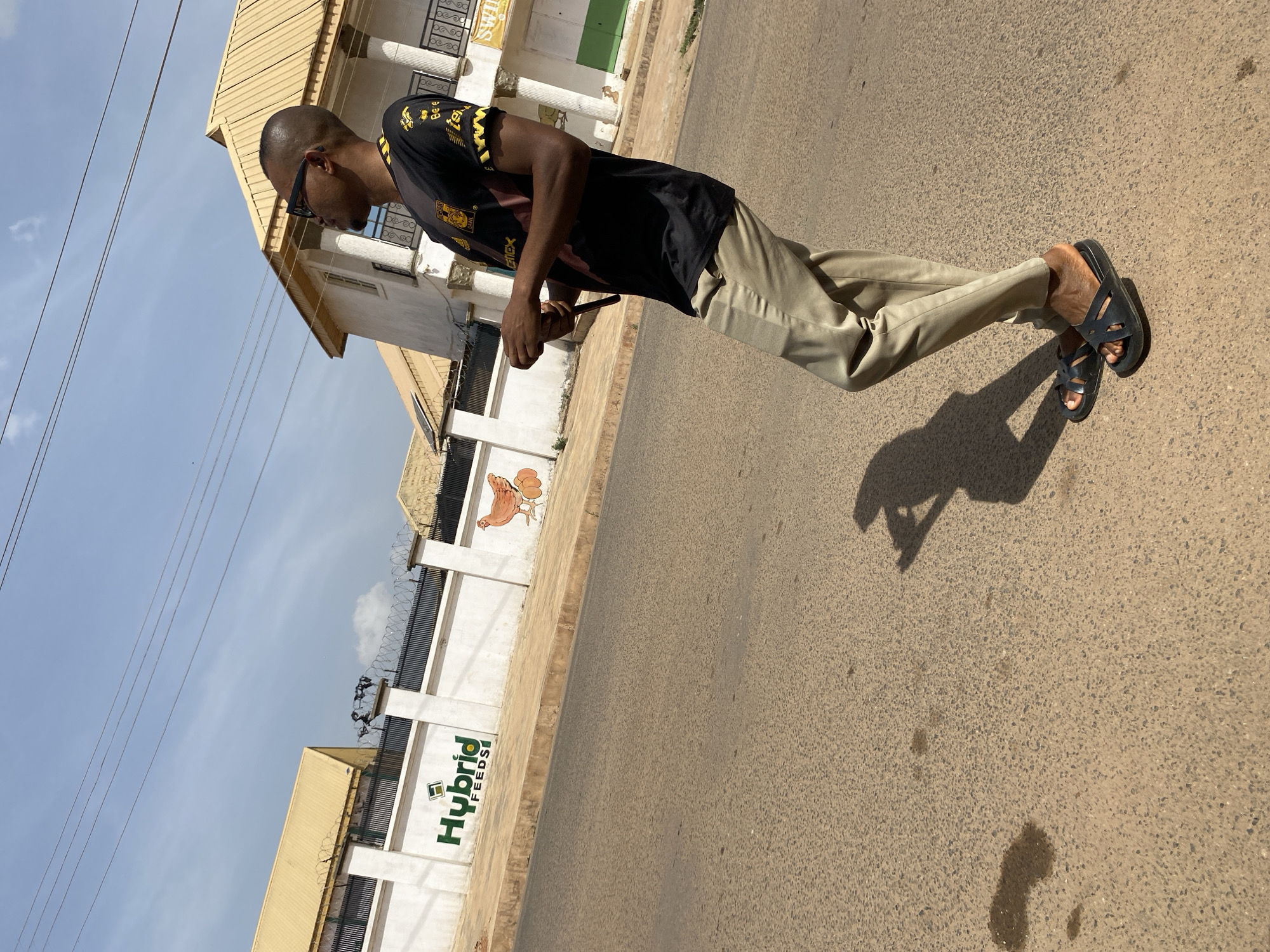 The image depicts a man standing on an asphalt road under bright daylight, likely in Jos, Nigeria. The man, seen in side profile, has dark skin, a shaved head, and wears sunglasses. He is dressed in a black t-shirt with yellow designs and text, light tan/beige trousers, and black sandals. He holds a dark object, possibly a phone, in his hands. His distinct shadow is cast on the ground to his right, indicating strong overhead sunlight.
The background features a white wall or fence topped with barbed wire, running parallel to the road. Prominently displayed on this wall is the green and white logo and text "Hybrid FEEDS," alongside a painted mural of a brown chicken and eggs. Behind the fence, several buildings with light yellow/gold roofs and white-painted structures are visible, consistent with local architecture. The sky above is mostly clear with a few thin power lines stretching across the upper left of the frame.
Notable visible text includes "Hybrid FEEDS" on the wall, and partial text like "Bee Len" and "LENEX" on the man's t-shirt. A distant sign on a building appears to begin with "SWI". The setting suggests an urban or semi-urban outdoor environment with clear, sunny weather.