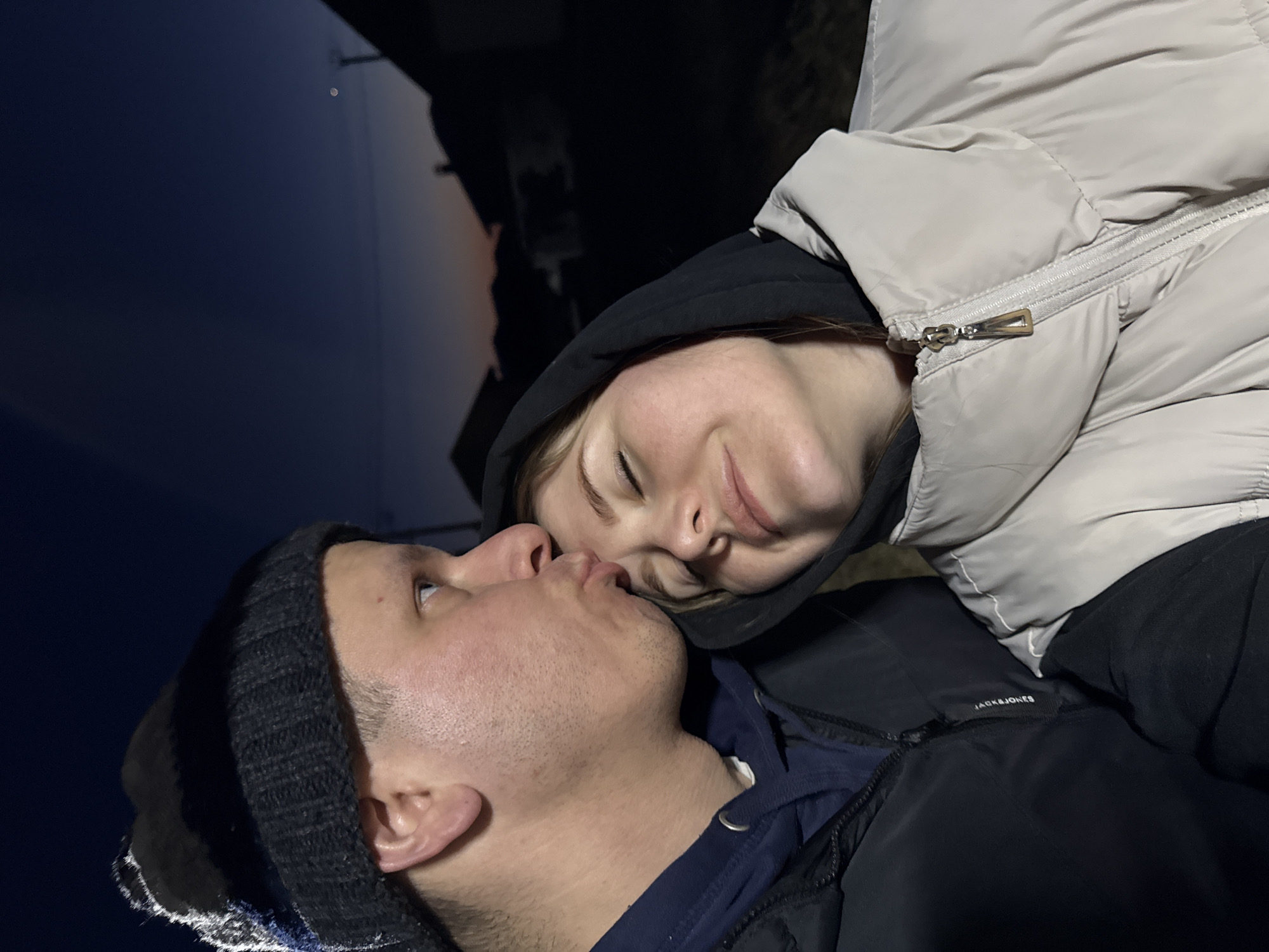 A couple is captured in a close-up, seemingly affectionate pose outdoors. The man, wearing a black knit hat, is positioned with his face looking upwards, while the woman, dressed in a light-colored, puffy jacket with a black hood pulled up, is angled downwards towards him. Their faces are close, appearing as if they are about to kiss or are in a moment of intimate connection. The background reveals a dark, indistinct cityscape possibly at dusk or night. The jacket has the logo "JACK&JONES" on it. There is a single light point above the couple. This photo may be taken in Gornyi (Dvurechenskoe S/P), Russia.