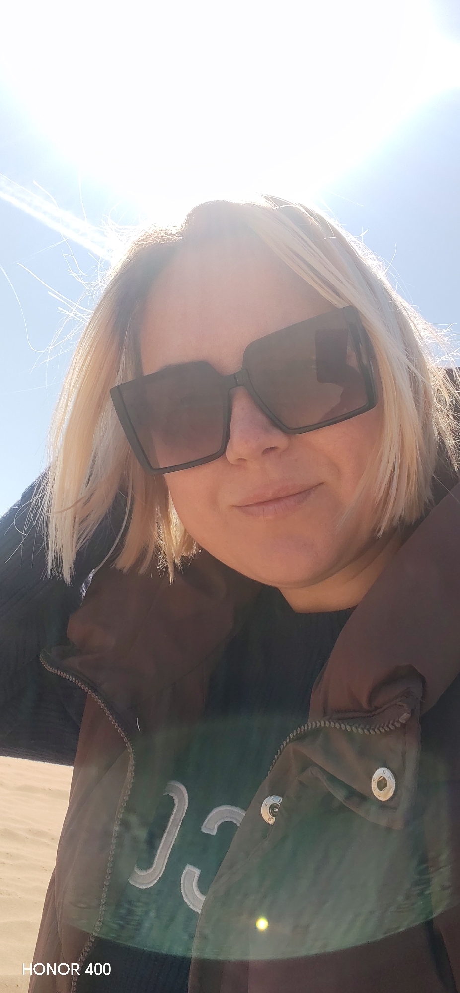 A woman with blonde hair and large black sunglasses is facing the camera. She is wearing a dark top and a brown vest. The sky is bright and sunny. The photo may have been taken outside in Noviy, Russia, though further information would be needed to confirm the location. The lower left corner contains the text "HONOR 400".