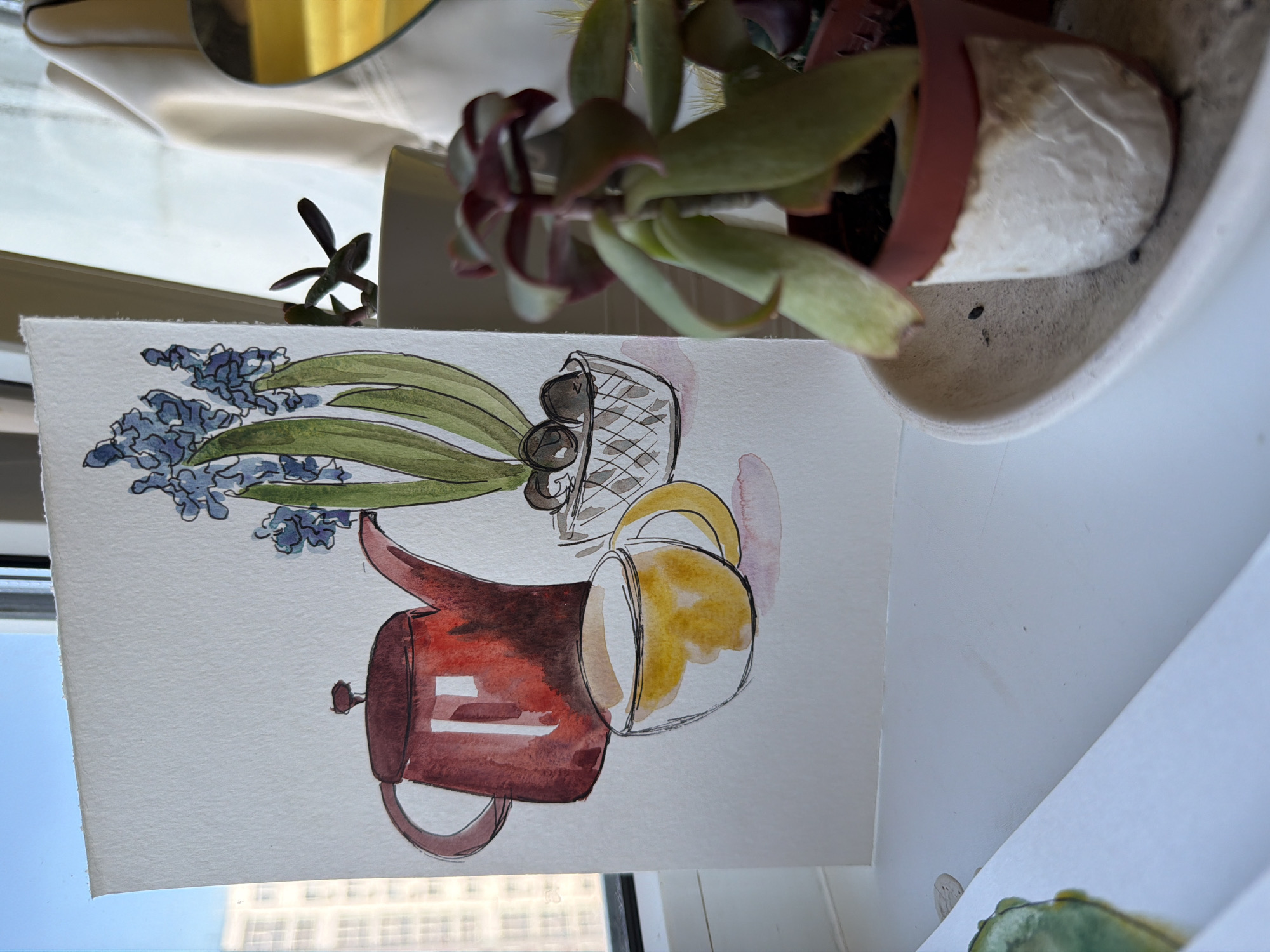A watercolor painting depicts a red teapot and a yellow teacup next to a pot of blue hyacinths. The painting is placed on a white windowsill. To the right of the painting is a potted succulent plant, wrapped in white textured paper. The scene is lit by natural light, suggesting it is daytime. In the background, out of focus, are buildings visible through a window, indicating an urban environment. The location is Ufa, Russia.