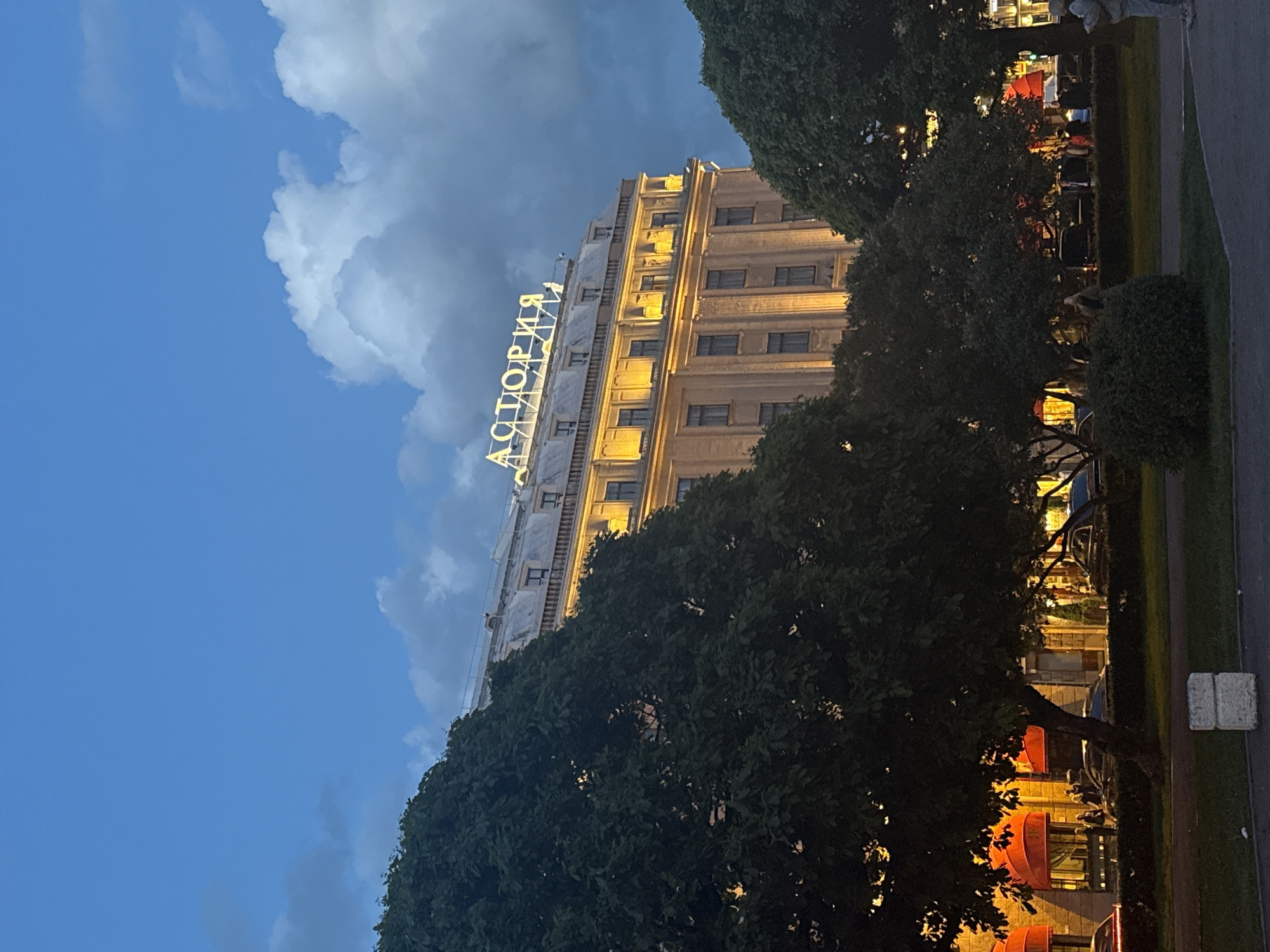 The image captures an evening scene in Saint Petersburg, Russia, featuring the grand Hotel Astoria. The sky is a deep blue, transitioning from twilight, with prominent white and grey cumulus clouds.
The Hotel Astoria building dominates the upper half of the frame (when oriented correctly), showcasing its classical architecture with multiple stories, numerous windows, and ornate detailing. The building is brightly illuminated by warm yellow lights, highlighting its facade. A large, prominent sign on the top of the building spells "АСТОРИЯ" (Astoria) in glowing white Cyrillic letters.
In the foreground and partially framing the building, there are dark green trees with dense foliage. Beneath these trees, to the right of the building, an outdoor area is visible. This area appears to be an outdoor cafe or restaurant, lit by warm artificial lights. Red awnings or umbrellas provide cover for tables where several people are seated, engaged in dining or socializing. A strip of well-maintained green grass borders a paved path or road, and a white rectangular concrete block is visible on the ground to the far right.
The time of day is clearly dusk or early evening, indicated by the diminishing natural light in the sky and the active artificial illumination of the building and outdoor spaces. The weather seems clear with distinct cloud formations.