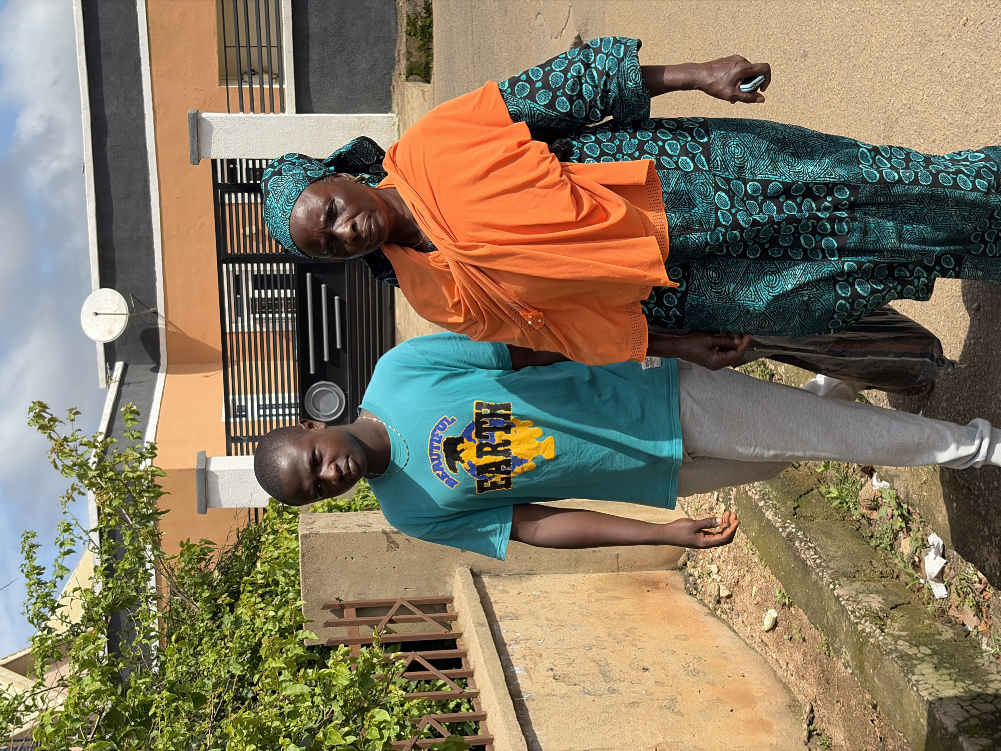 A young man and an older woman walk side-by-side down a paved street in Jos, Nigeria. The man, on the left, wears a teal t-shirt with the words "Beautiful Earth" printed on it and light gray sweatpants. He has a necklace on. The woman, on the right, wears a teal and black patterned dress, a teal headscarf, and an orange shawl draped over her shoulders. She is holding a cell phone in her right hand. Behind them, a concrete wall and a metal fence line the street, with lush green foliage growing behind the fence. The background features a modern building with orange and gray accents, and a satellite dish mounted on the roof. The sky above is partly cloudy.