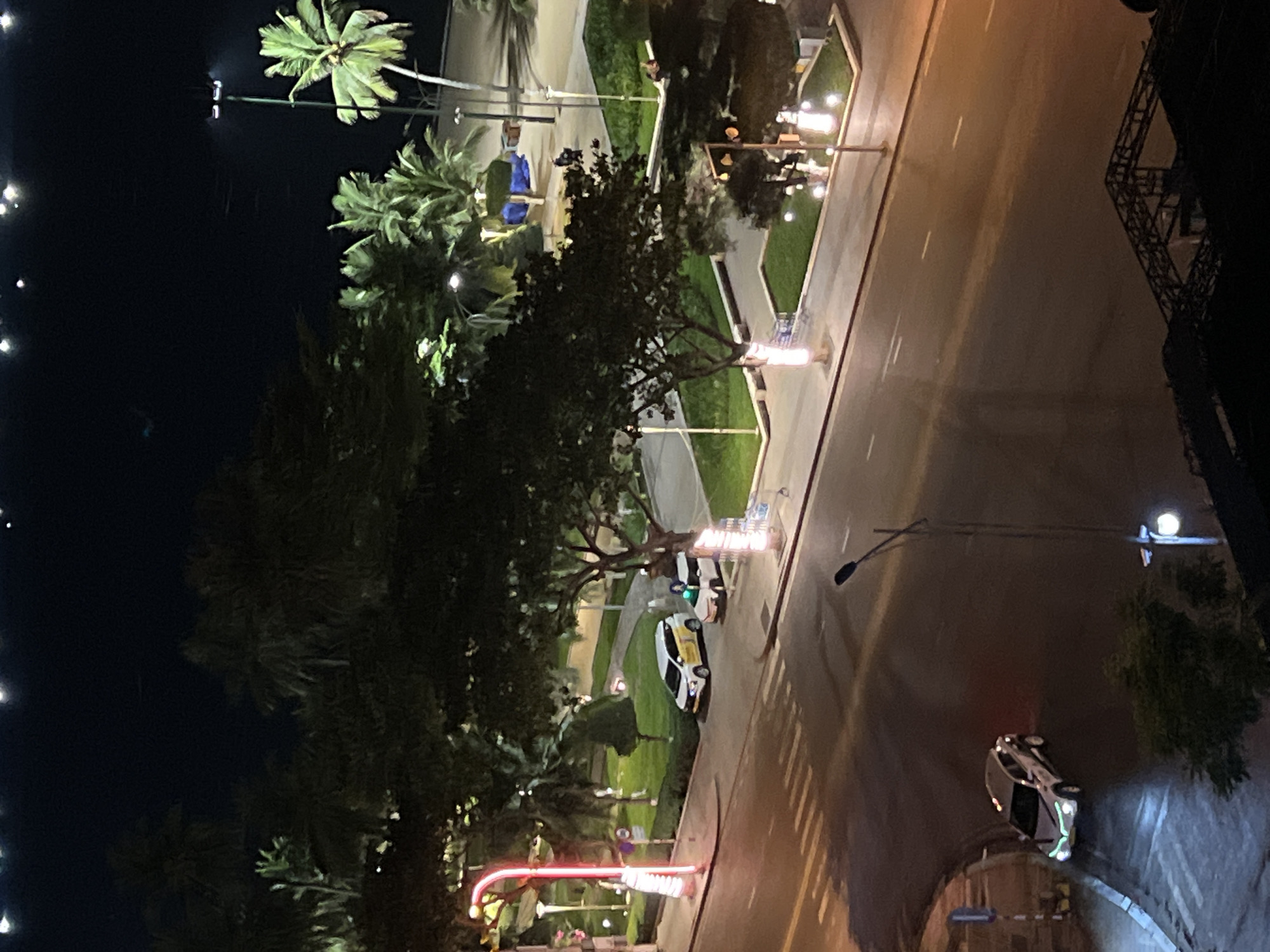 This aerial nighttime photograph captures a street scene in Nha Trang, Vietnam, likely near a coastal area given the presence of palm trees. The image is dominated by the darkness of night, punctuated by artificial lighting from streetlamps, buildings, and vehicles.
In the foreground, a paved road curves to the left, marked with white dashed lines indicating lanes. A silver sedan is parked on the left side of the road, its headlights and front lights illuminated. Across the street, a manicured green area with scattered trees and bushes is visible.
Further back, two white vehicles, one a minivan and another appearing to be a car, are parked on the side of the road. A tall, red, illuminated signpost stands on the left side of this area. Beyond the parked vehicles, more palm trees and other lush vegetation are present. In the distance, glimpses of what appears to be a beach or sandy area can be seen, along with streetlights. The sky is dark with a few scattered, bright points of light, possibly distant stars or decorative lighting. The overall impression is a relatively tranquil urban environment at night, with a tropical atmosphere.