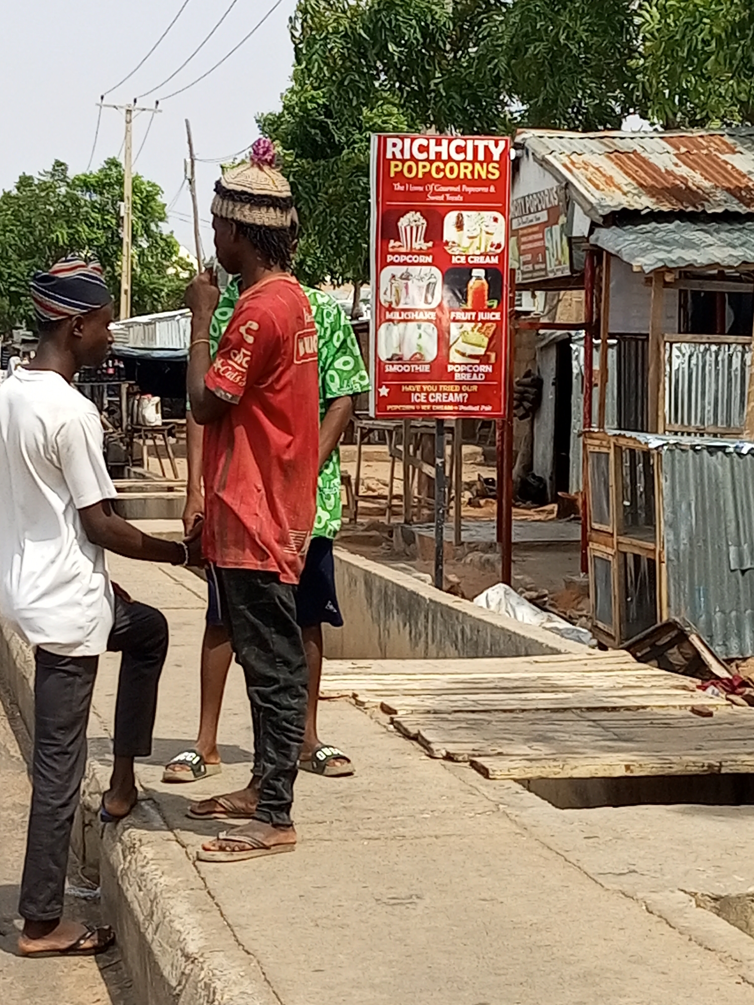 In an outdoor urban setting, possibly near a roadside market, three young males are the central figures. The individual on the far left wears a white t-shirt and black pants, with a striped cap. Next to him, a young man in a red t-shirt and black pants wears a distinctive knitted cap adorned with a pink pompom. A third male, partially visible in a green t-shirt with a white pattern, stands to the right. They appear to be interacting, with two of them holding hands or gesturing. In the background, a red and white sign advertising "RICH CITY POPCORNS" is prominently displayed, showcasing images of popcorn, ice cream, milkshakes, fruit juice, smoothies, and popcorn bread. Adjacent to the sign is a rustic stall with a corrugated metal roof and wooden shelves. The ground is a mix of concrete and wooden planks, suggesting a somewhat uneven or constructed walkway. Electrical wires are visible overhead, and lush green trees form a backdrop. The scene is brightly lit, indicating daylight.