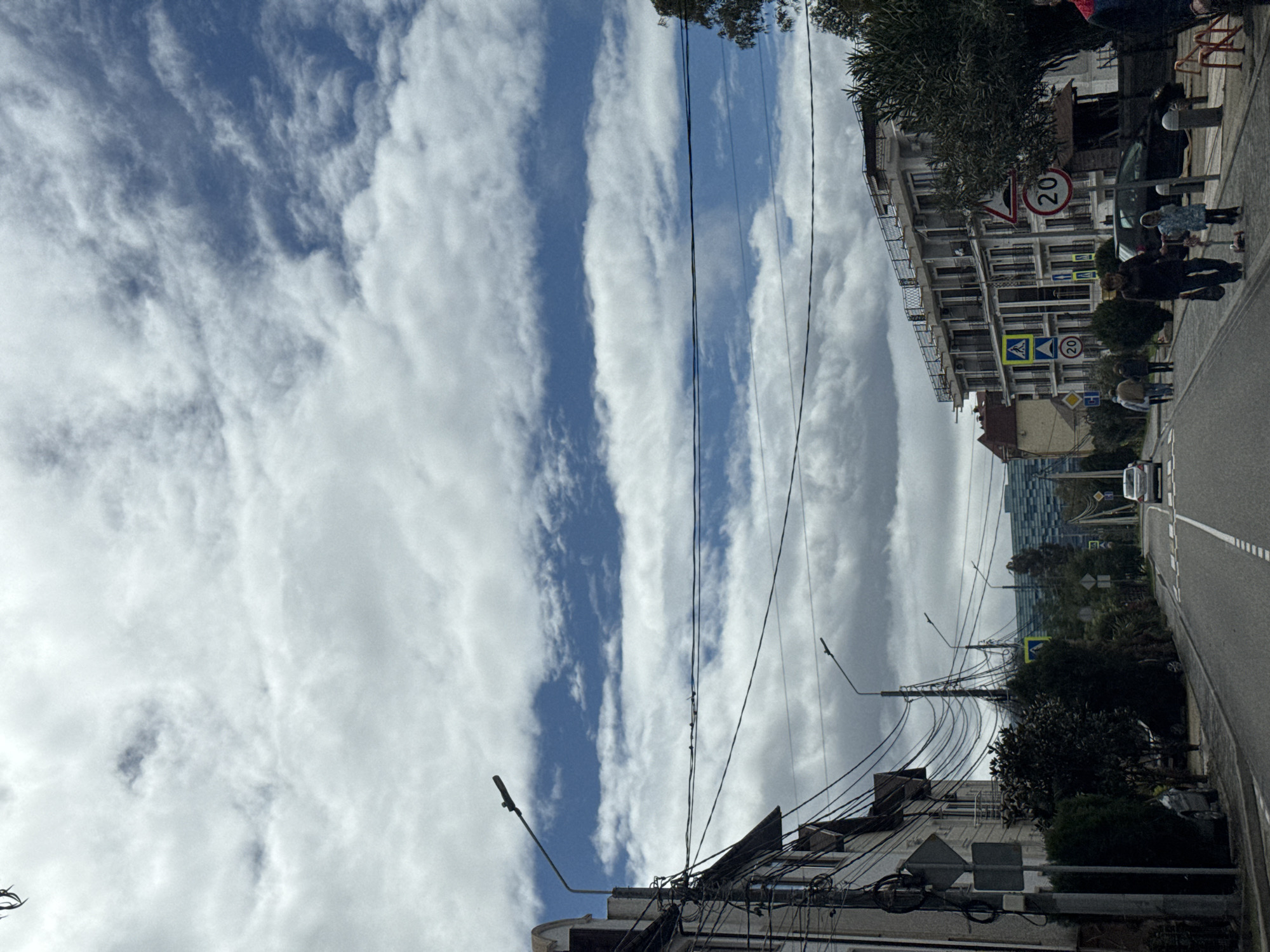 The image shows a street scene in Sochi, Russia, under a sky with striped cloud formations. The street has a pedestrian crossing. People are walking on the sidewalk next to buildings. The buildings appear to be mixed-use, including apartments and shops. A car is on the road. Street signs are present, including speed limit signs. Overhead power lines are visible.