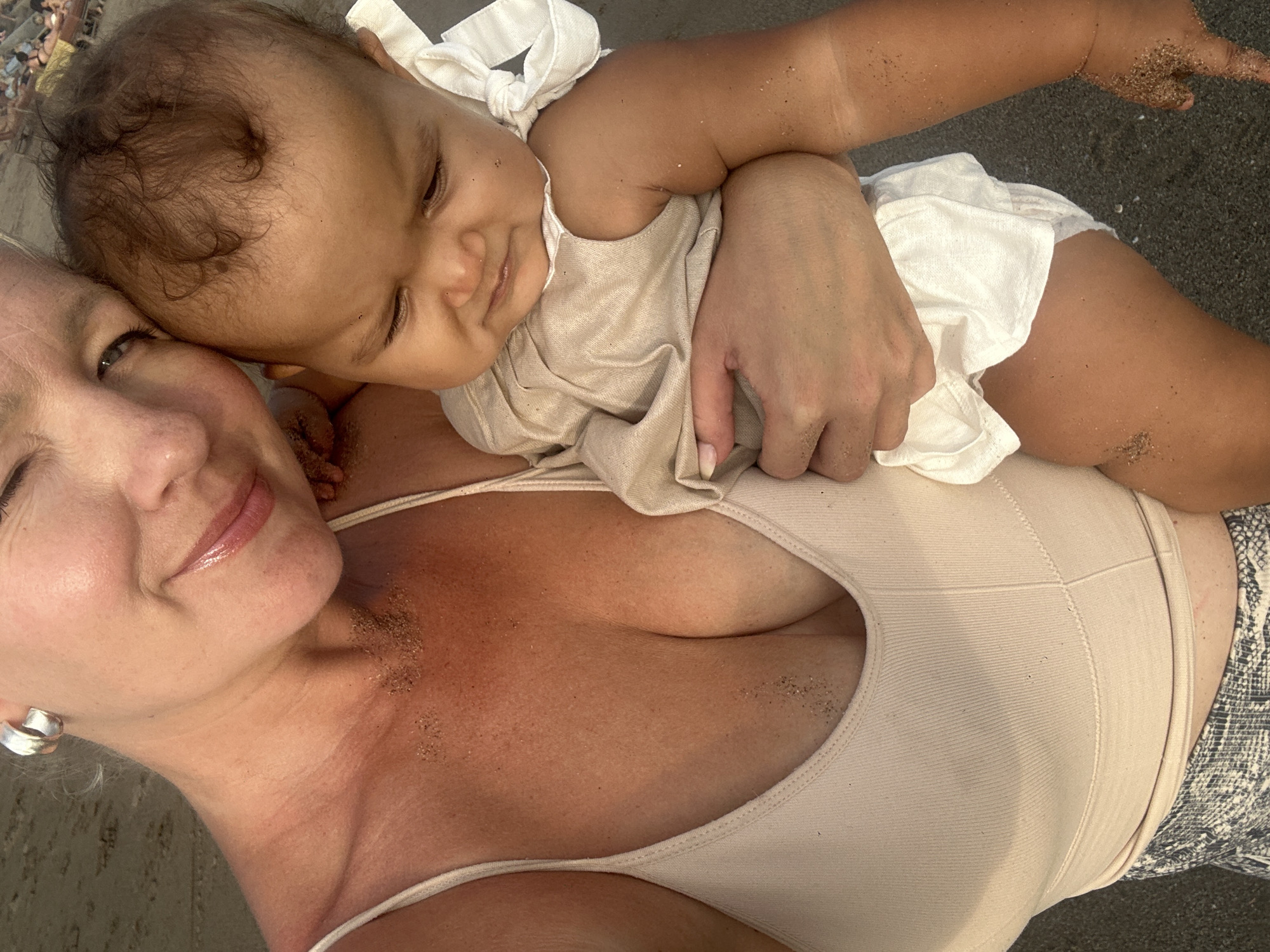 An adult woman is holding a baby on a beach in North Kuta, Indonesia. The woman is wearing a beige tank top and a snake-print skirt, and the baby is dressed in a light-colored outfit. Both are covered in sand. The background shows more beach with people in the distance.