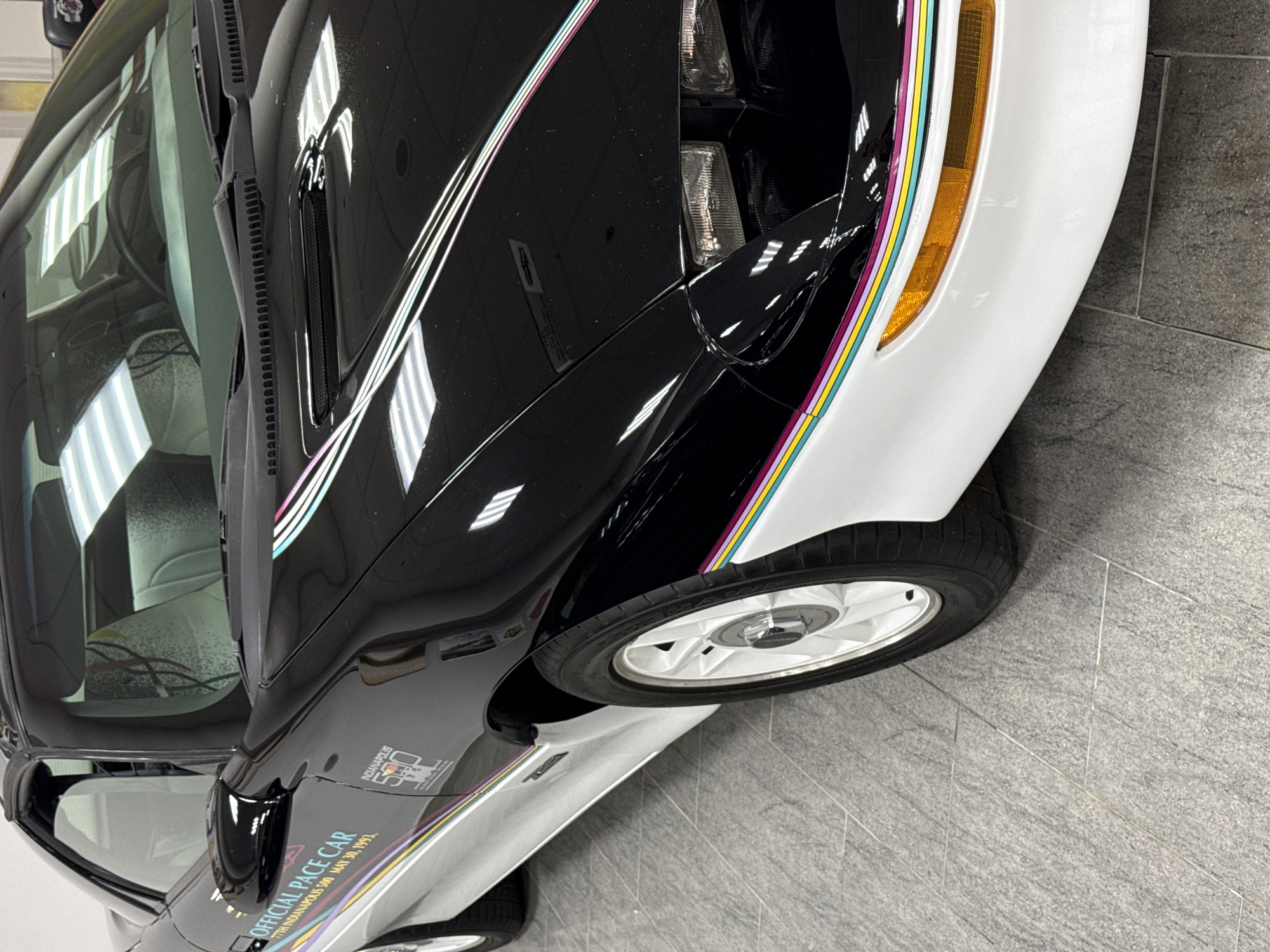 An eye-level, slightly angled shot captures the front left and side of a well-maintained car, likely in a showroom or private collection in Sirius, Russia. The vehicle is a classic "Official Pace Car," featuring a distinctive two-tone paint scheme: glossy black on the upper body, including the hood and fender, transitioning to white on the lower body and front bumper. A vibrant set of thin, parallel stripes in pink, light blue, yellow, and purple delineates the color change, running along the fender and side.
The front left amber turn signal and part of the headlight assembly are visible, contrasting with the white bumper. A white multi-spoke wheel with a central hub cap is fitted on the front. Reflective surfaces of the car's body clearly show the overhead fluorescent lighting of the indoor environment. A side mirror is present, reflecting part of the interior.
Prominently displayed on the side of the car, near the door, is the text "OFFICIAL PACE CAR" in capital letters. Below this, further details read "77TH INDIANAPOLIS 500 MAY 30, 1993." An additional logo for "INDIANAPOLIS 500" with a checkered flag motif is also visible above the main text. The interior of the car, including parts of the seats and dashboard, is visible through the windshield. The car rests on a grey, textured tiled floor. No activity is taking place; the car appears to be on static display.