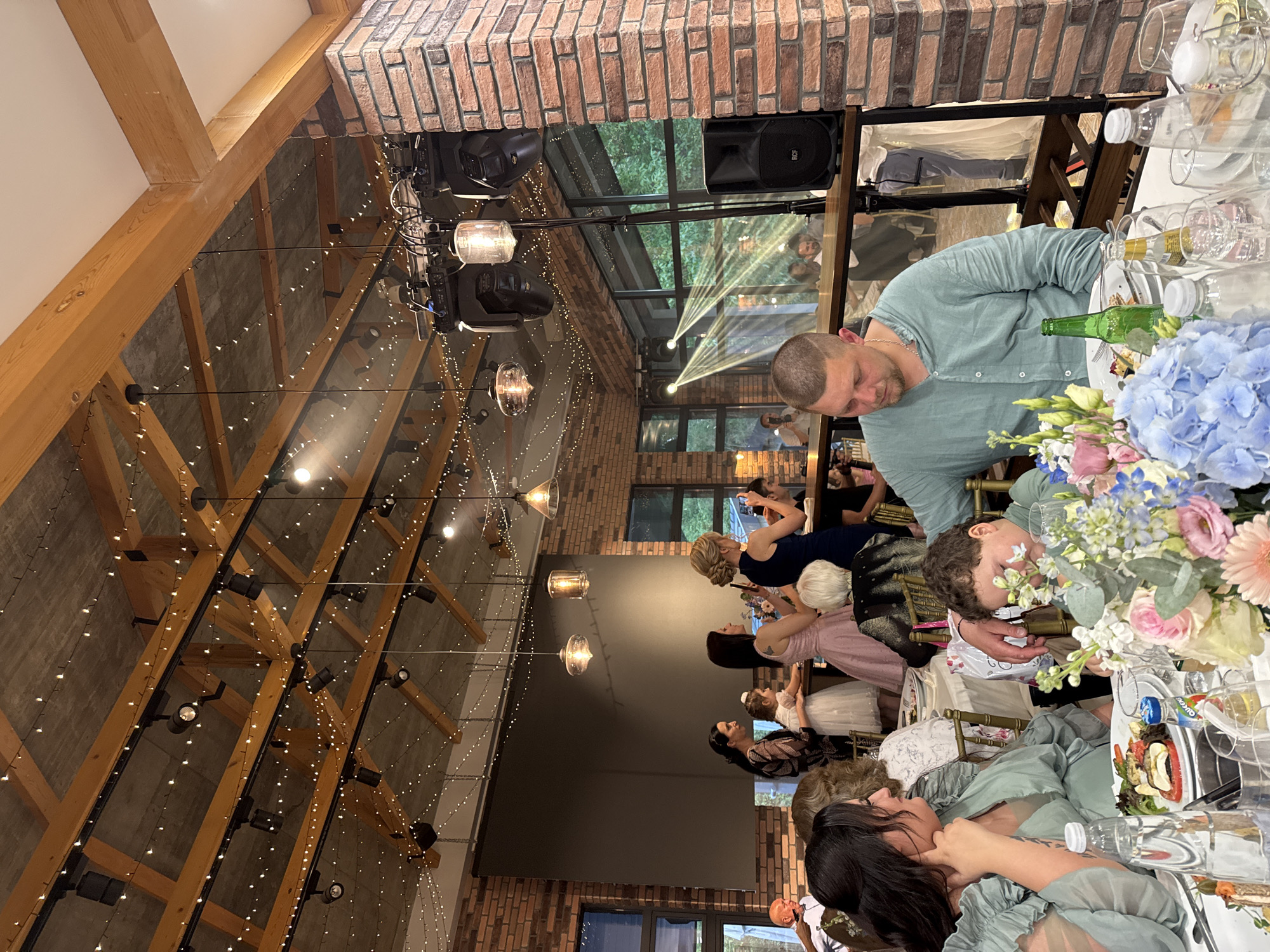 The image depicts an indoor event, possibly a wedding reception, in what appears to be a renovated industrial building with exposed brick walls and a high ceiling featuring wooden beams and string lights. People are seated at tables, engaged in conversation. A man in a light green shirt is seen near a table with floral arrangements and glassware. A child is also at the table, seemingly interacting with the flowers. Other individuals are in the background, some using phones, indicating they might be taking photographs. The building has a glass wall through which trees are visible. The location is possibly in or near Mogilovo, Bulgaria. A sound system is set up, suggesting music or speeches are part of the event.