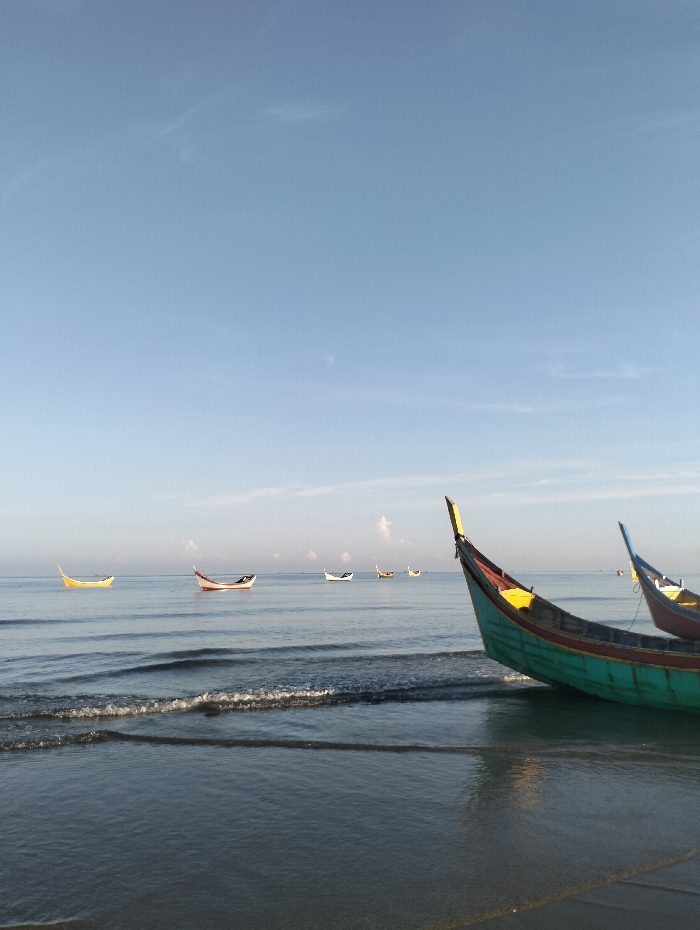 A serene beach scene under a clear blue sky features several traditional fishing boats. In the foreground, a vibrant green and yellow boat rests on the wet sand. Further out, smaller boats dot the calm ocean waters, their colorful hulls reflecting in the tranquil surface. Gentle waves lap at the shoreline, creating ripples that mirror the expansive sky above. The image evokes a sense of peace and simplicity.