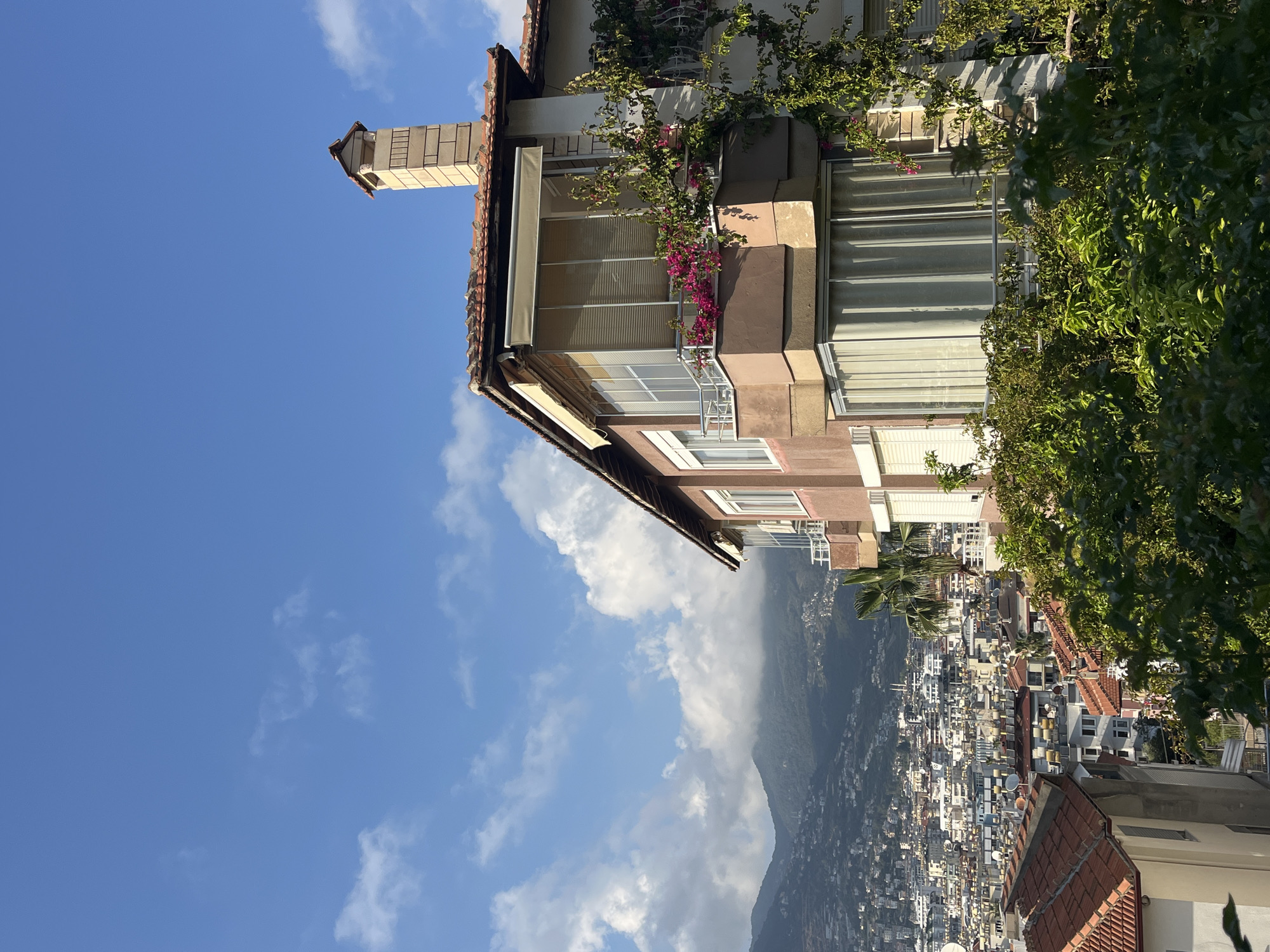 This image, when viewed in its correct orientation (rotated 90 degrees counter-clockwise from the upload), captures a sunny daytime scene of a coastal city and mountains.
In the foreground, on the right side, stands a multi-story building, likely an apartment complex, featuring light-colored facades and a terracotta-tiled roof. A decorative chimney structure extends upwards from the rooftop. Several balconies are visible, adorned with lush green foliage and vibrant pink bougainvillea flowers. Some windows have blinds or awnings for shade.
Beyond the foreground building, a sprawling cityscape fills the midground, stretching across a hillside. The city consists of numerous white and light-colored buildings with red-tiled roofs, characteristic of Mediterranean architecture. Many rooftops feature satellite dishes. The urban area extends towards a large mountain range that dominates the background, with some peaks partially enveloped by soft white clouds.
The sky above is a brilliant clear blue, interspersed with fluffy white cumulus clouds. Bright sunlight illuminates the scene, casting distinct shadows, indicating it is daytime, likely late morning or early afternoon. The setting portrays a vibrant city integrated with natural landscapes, consistent with the location of Alanya, Turkey. There is no visible text in the image.