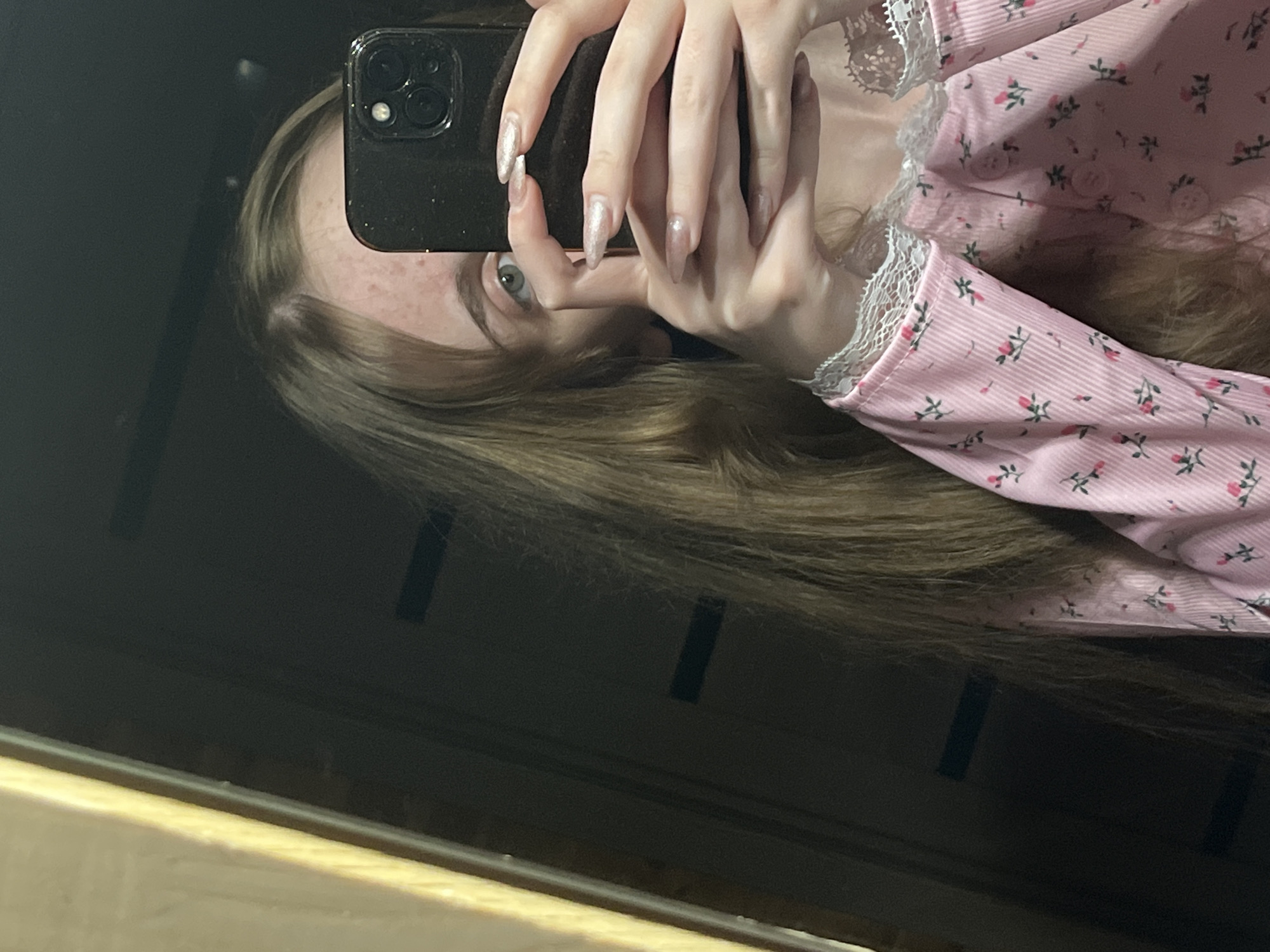 A young woman with long, light brown hair takes a selfie in a mirror. She is wearing a pink pajama top with a floral pattern and white lace trim. Her right eye is visible, along with her hands holding a black smartphone. The background is dark and out of focus, with a reflection of what appears to be a doorway or closet. The lighting is dim, creating a soft and intimate atmosphere. The image was captured on November 23, 2022, at 23:17 in Ashkadar, Russia.