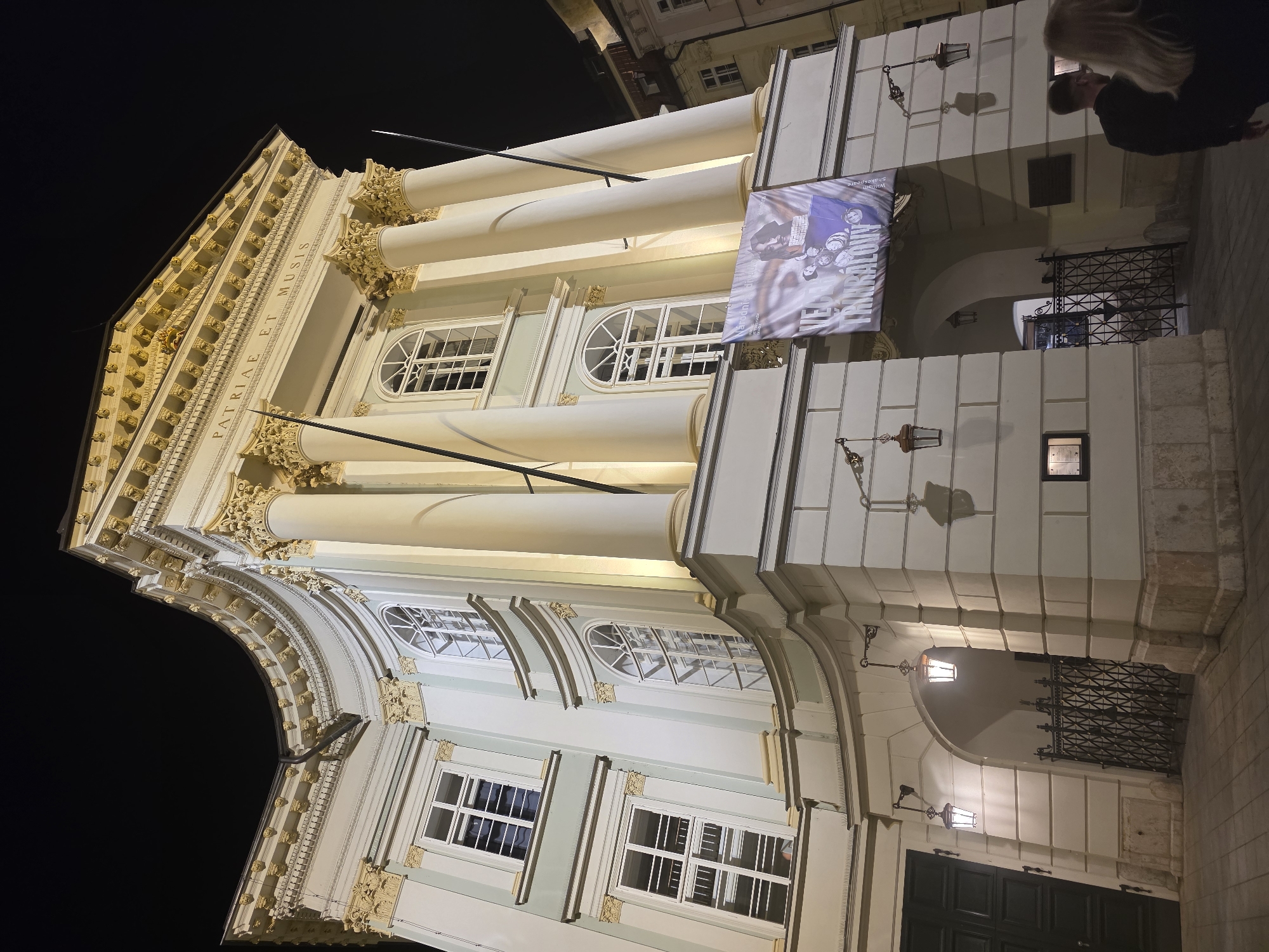 The image depicts the Estates Theatre (Stavovské divadlo) in Prague, Czech Republic, at night. The multi-story building features a curved facade illuminated by artificial light, set against a dark, featureless sky. The architecture is grand, characterized by a central section with four prominent columns supporting a pediment. The pediment bears the Latin inscription "PATRIAE ET MUSIS" (To the Homeland and the Muses) in capitalized gold lettering.
The building's facade is a light, possibly pale green or off-white color, accented with intricate golden or yellow architectural detailing, including ornate column capitals (likely Corinthian style), cornices, and window surrounds. Windows on the curved section are arched on the lower level and rectangular above, while windows on the flatter left section are all rectangular. All visible windows have white frames.
Below the central columns and pediment, a large, vertically hung banner is displayed. The banner is predominantly purple and white, featuring the text "NÁRODNÍ DIVADLO" (National Theatre) and "VĚČER TŘÍKRÁLOVÝ" (Twelfth Night), along with stylized images of figures, possibly actors.
At ground level, there is an arched entryway secured by an ornate black wrought-iron gate on the right, and a dark, possibly wooden, double door on the left. Two ornate wall-mounted lanterns, emitting a warm glow, are affixed near the arched entrance. The ground in front of the building is paved with grey interlocking pavers. In the lower right corner, partially visible from behind, are two individuals: a male with dark hair and a female with long light-colored hair, walking from right to left across the frame.