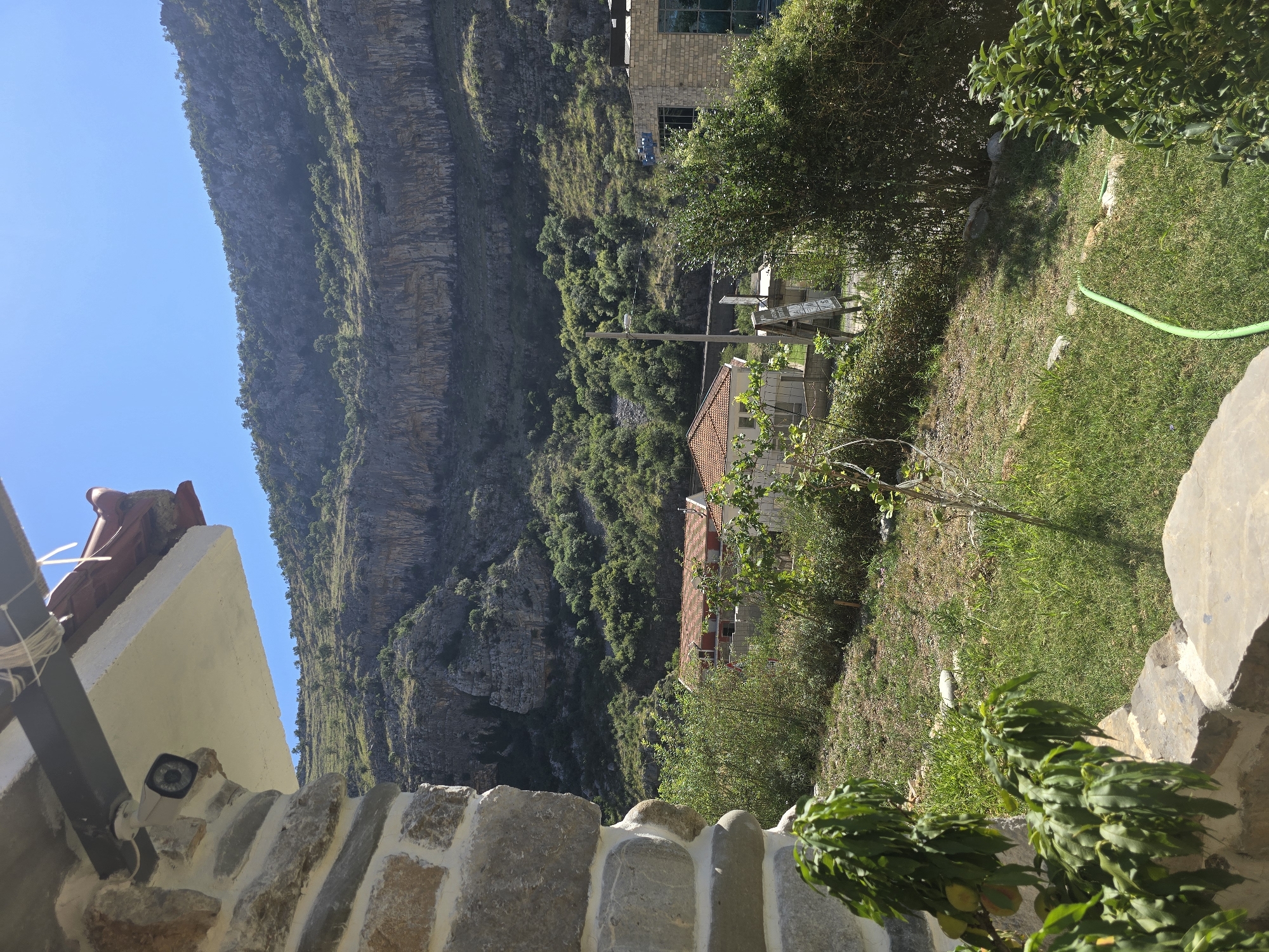 This is a landscape image taken from a slightly elevated position, looking out over a valley towards a large, rocky mountain range under a clear blue sky. In the foreground, to the left, is a stone wall and a white overhang with a security camera mounted on it. Below the overhang, there are green trees and foliage. In the middle ground, there are a couple of houses with orange tile roofs and some vegetation. Further into the background, the mountains rise steeply, covered in sparse green vegetation. The overall scene depicts a rural or mountainous area bathed in bright sunlight.