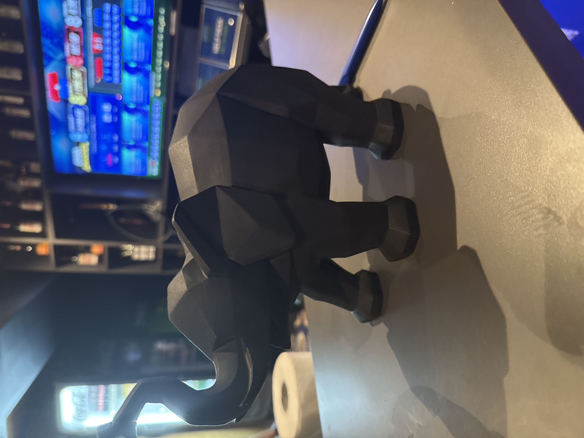 A low-angle shot depicts a dark gray, geometric-style elephant statue standing on a light gray surface, possibly a table or counter. In the background, partially out of focus, is a brightly lit beverage cooler and a digital display with brightly colored text. The environment appears to be a dimly lit interior space, potentially a bar or restaurant.