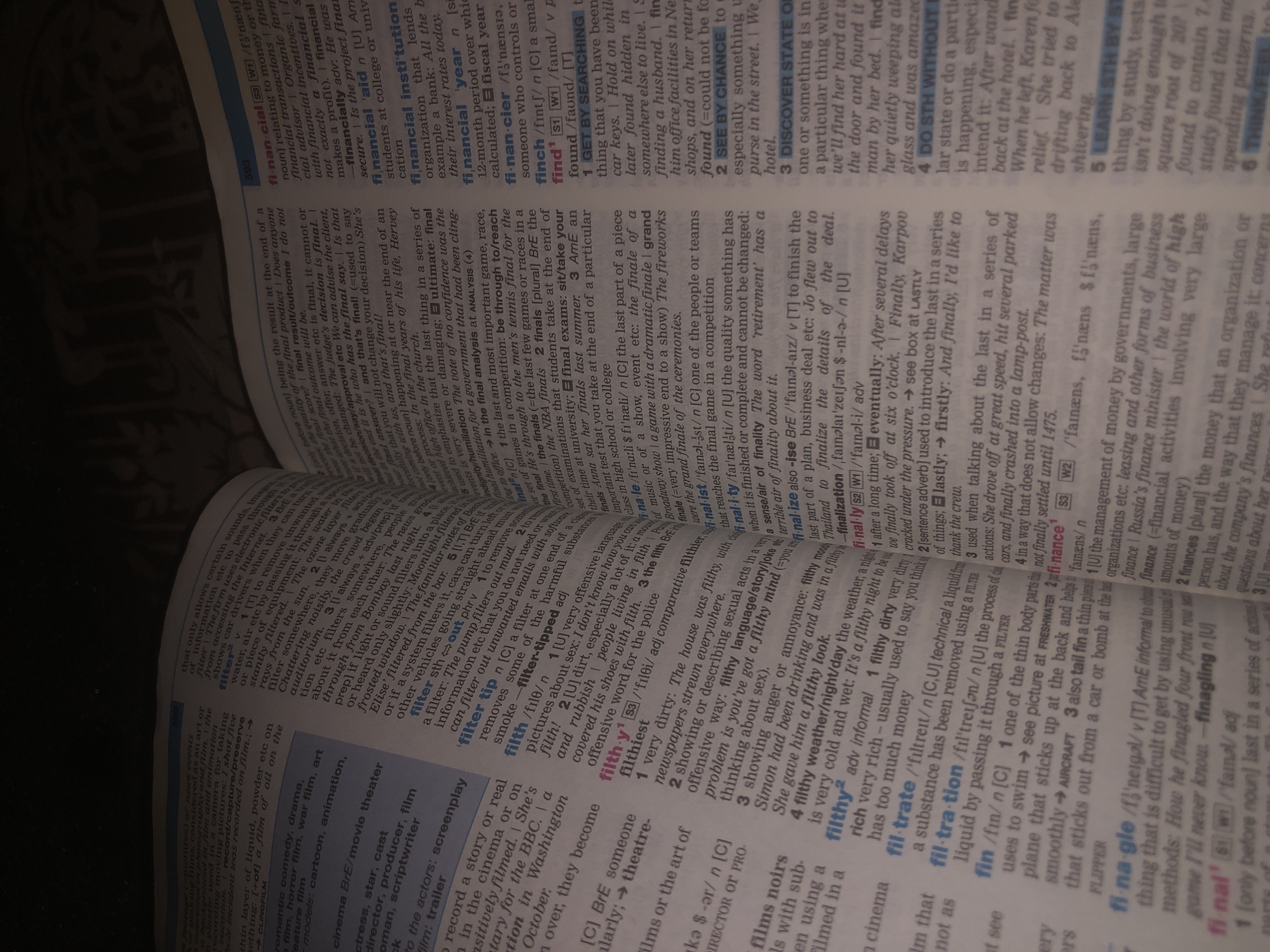The image shows a close-up view of an open book, likely a dictionary, lying flat on a surface. The pages are filled with dense text, featuring entries and definitions in English. The lighting appears to be artificial, suggesting an indoor setting, possibly at night or in a dimly lit room. The camera is angled downwards, focusing on the printed words and letter definitions.
The visible text includes entries such as "financier," "financial institution," "financial year," "finch," "find," "filter," "filth," and "finale." There are also phonetic transcriptions and grammatical information associated with each word. The text is predominantly black print on off-white paper, with some bolding and italicization used for emphasis and categorization. The book's binding is not visible, but the pages are slightly curved, indicating it's a substantial volume. The overall impression is one of study and learning, with the focus solely on the textual content of the book. No people, specific objects beyond the book itself, or environmental details like weather or time of day are discernible.