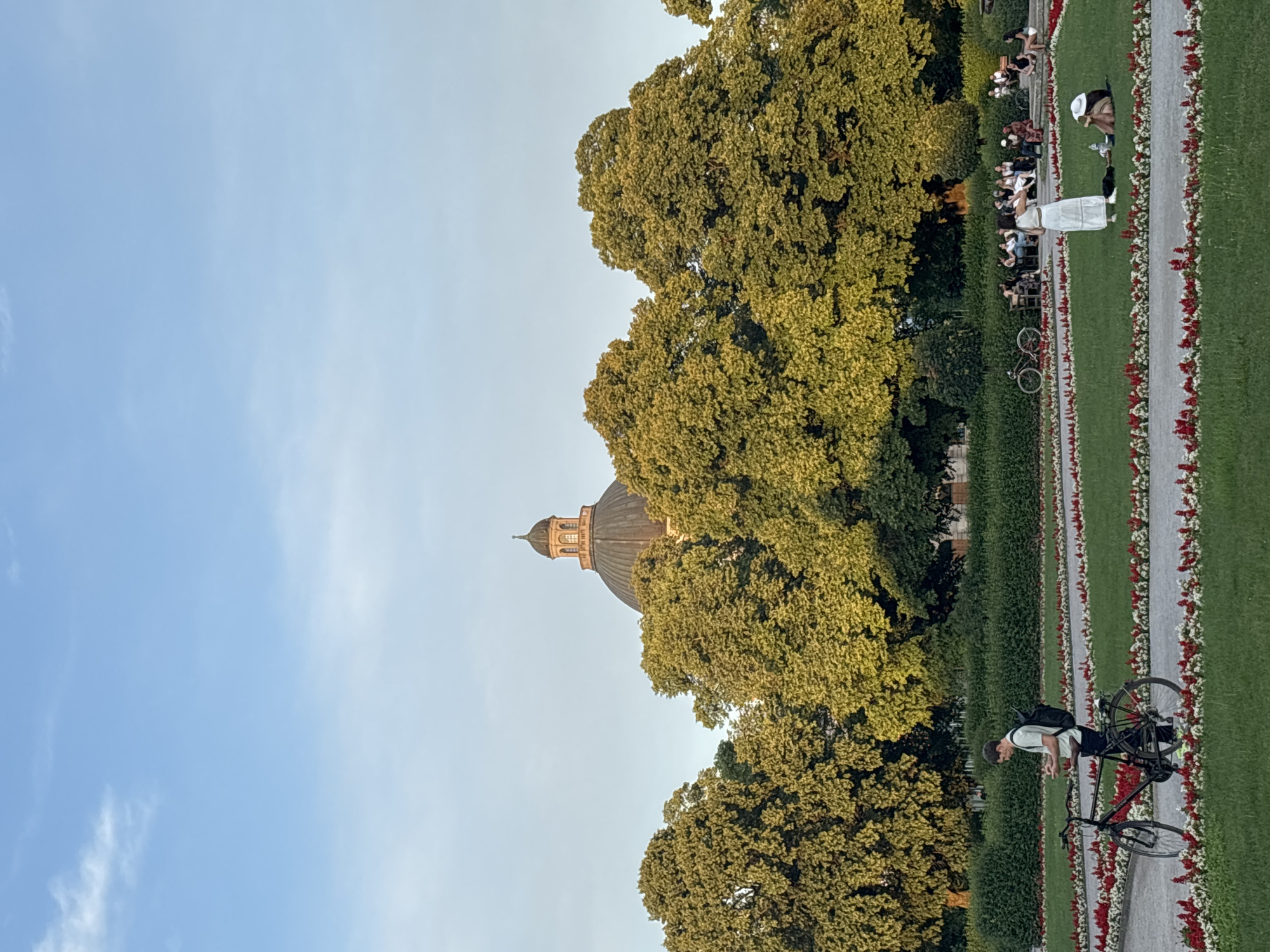 The image shows a park in Munich, Germany, with a large dome building in the background. The park features manicured lawns, flower beds, and trees. Several people are scattered throughout the park, some sitting and relaxing, while others are walking or cycling. In the foreground, a man with a backpack is standing next to his bicycle, looking at his phone. Further back, a group of people are gathered, some sitting on benches and others standing. The sky is clear and blue, with a few wispy clouds. The overall scene conveys a sense of peaceful leisure in an urban green space.