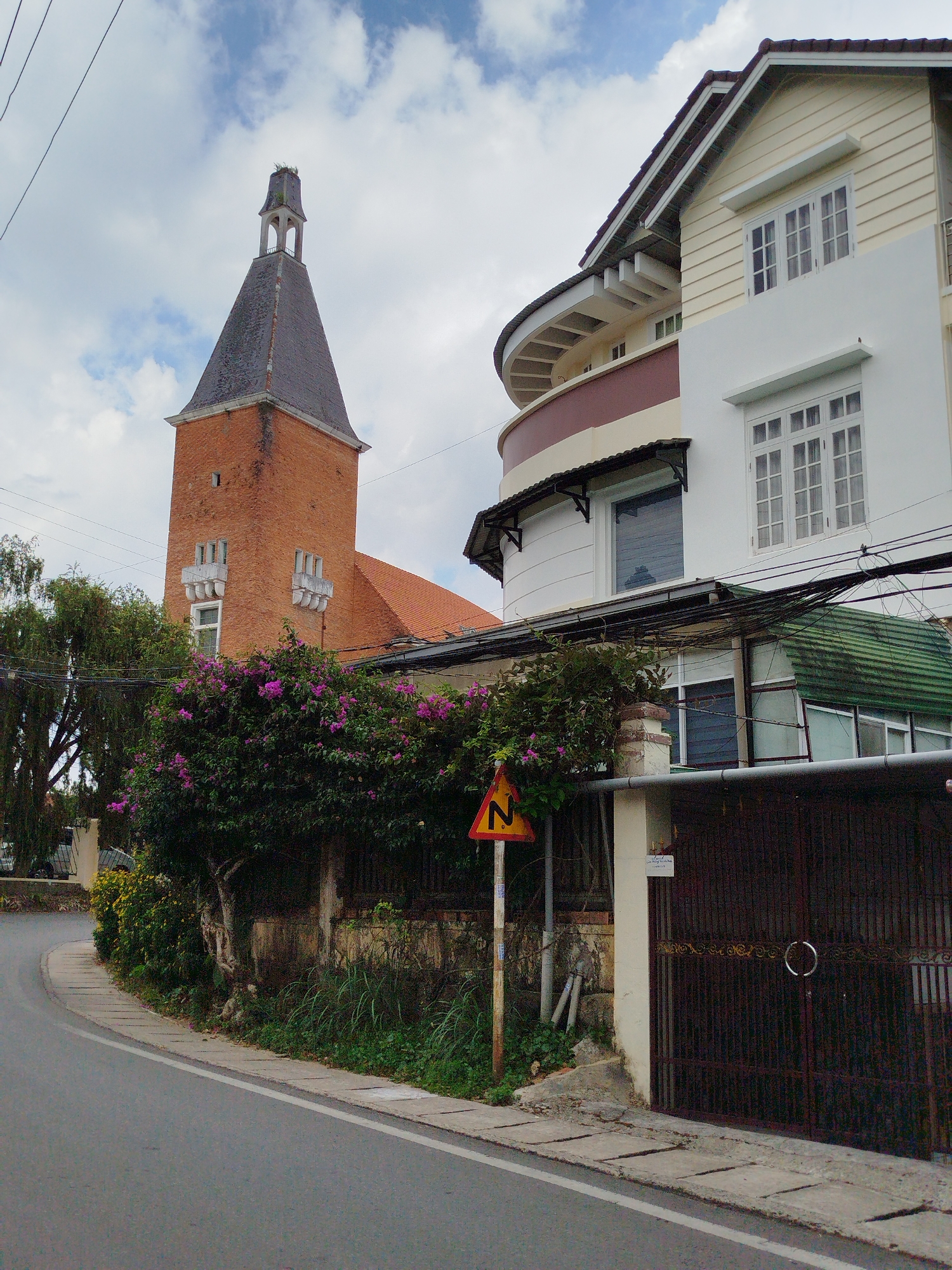 A street scene in Lâm Viên - Đà Lạt, Vietnam, features a red brick tower with a conical slate roof on the left and a multi-story house on the right. The house is a mix of white, beige, and maroon-painted sections with visible windows and curved architectural elements. A dark metal gate stands along the right side of the street behind a low stone wall topped with flowering bushes. The foreground shows a road with a solid white line along the edge, and a triangular road sign on a pole is visible behind the flowering bushes. Power lines are strung overhead. The sky is overcast with scattered clouds.