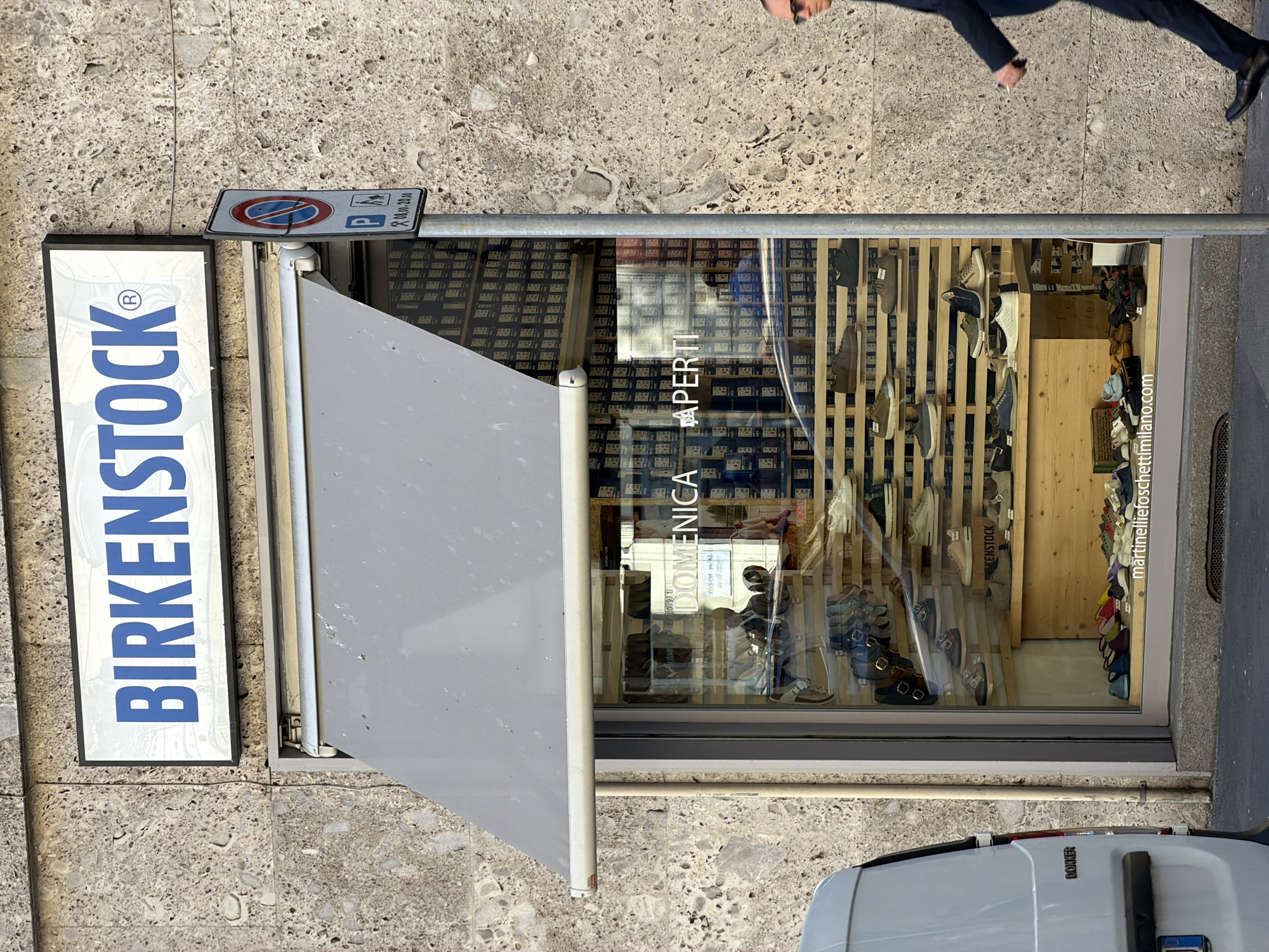 The image shows the exterior of a Birkenstock store in Milan, Italy, as indicated by "martineliefoschettimilano.com" visible in the window. The store has a prominent Birkenstock sign and a partially extended gray awning. The storefront window displays a variety of Birkenstock shoes arranged on wooden shelves. A sign in the window reads "Domenica Aperti," indicating the store is open on Sundays. A no-parking sign is mounted above the awning. A white vehicle is partially visible at the bottom of the frame, and a person in dark clothing is walking on the sidewalk above the store. The street and sidewalk appear to be constructed of stone or concrete.