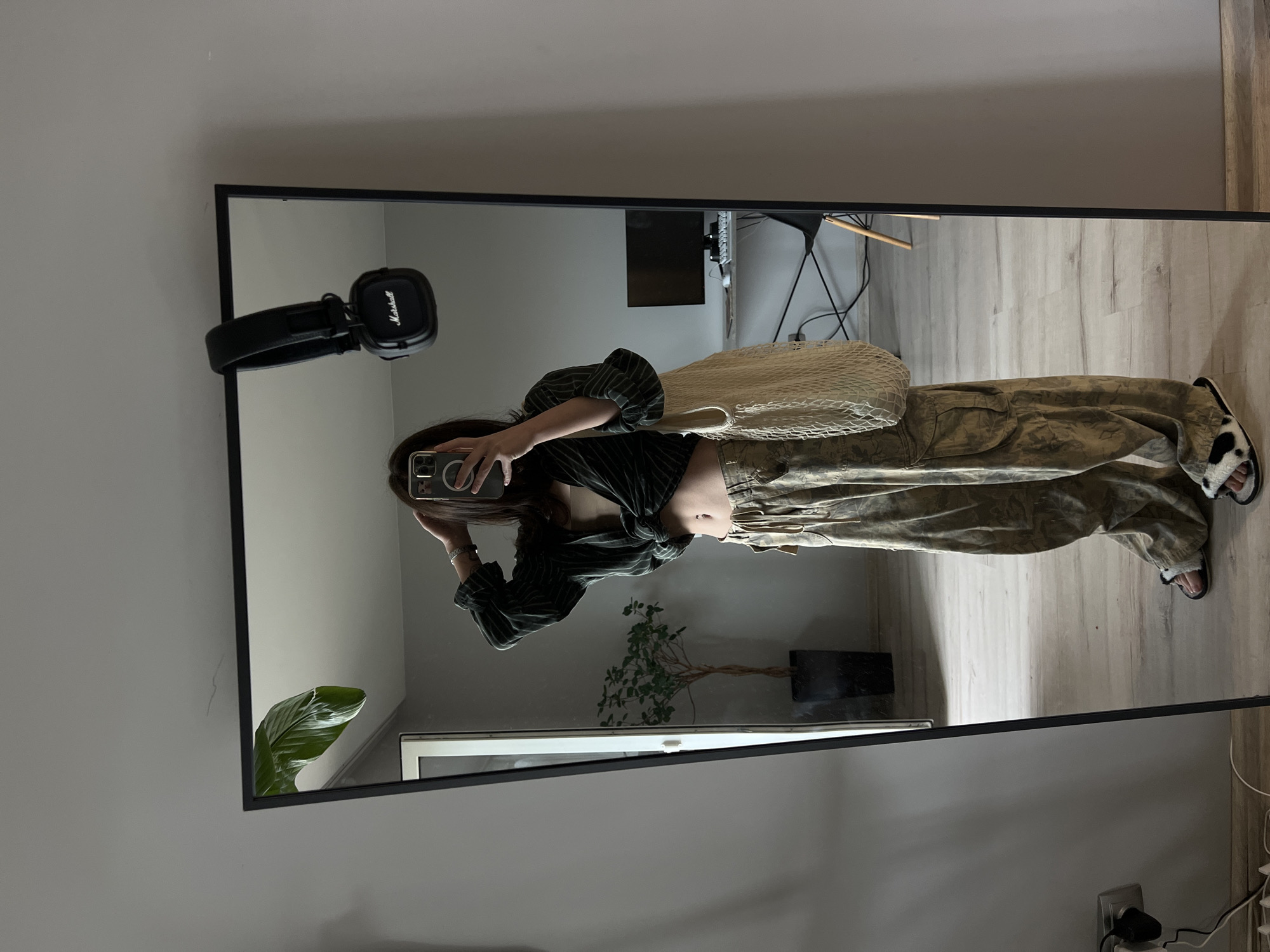 A full-body mirror selfie is captured in a well-lit room, likely a bedroom or dressing area, with gray walls and light wood-effect flooring. The subject, a person with dark hair, is standing and looking into the mirror, holding a smartphone in their right hand to take the picture. They are wearing a dark, striped, unbuttoned shirt tied at the waist, revealing a nude-colored camisole and their midriff. Their pants are loose-fitting cargo pants with a camouflage-like pattern in shades of green and brown. On their feet, they wear black slide sandals with white fluffy accents and a black and white cow-print pattern. A beige mesh tote bag is slung over their left shoulder. A pair of black Marshall headphones is hanging from the top edge of the mirror. In the reflection, a large potted plant with broad green leaves is visible to the left, and a desk with a computer monitor, keyboard, and chair is situated in the background on the right. The overall impression is casual and stylish. No specific landmarks or indicators suggest the location is Ternopil, Ukraine.