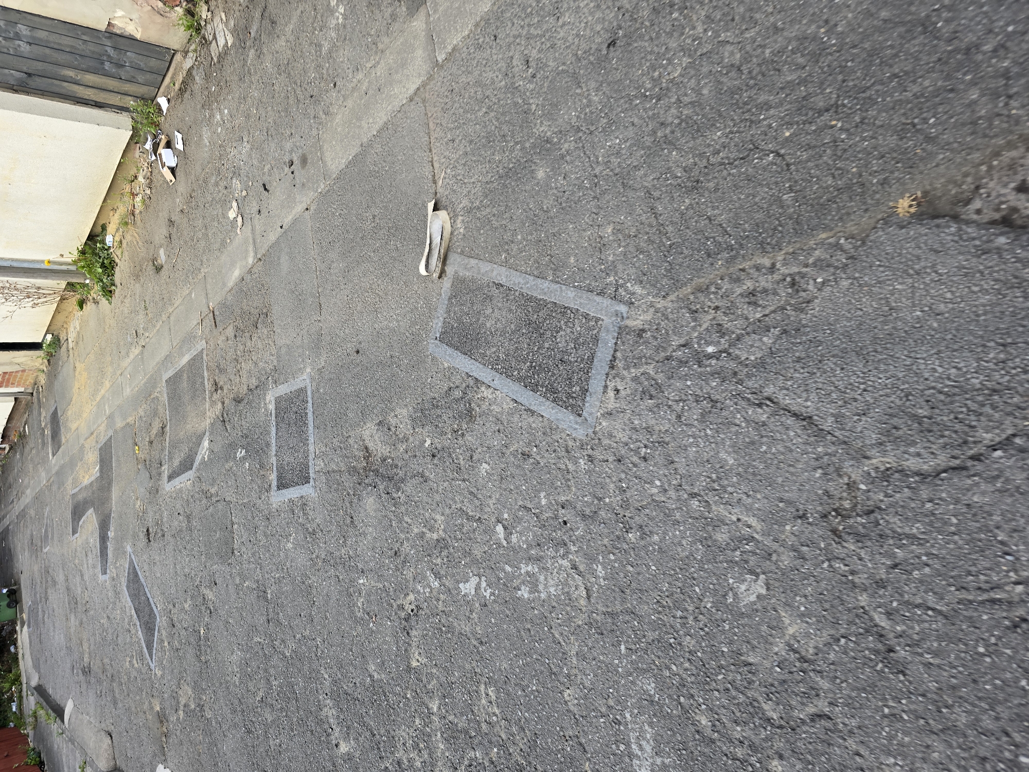 The image shows an asphalted alleyway in Stockton-on-Tees, United Kingdom, featuring multiple rectangular metal frames embedded in the pavement. These frames appear to be access covers or grates, some of which are filled with dark material, possibly asphalt. The alley is lined on the right by a cream-colored building with a dark wooden door and on the left by a low concrete curb. There is debris scattered along the side of the building, including crumpled paper and cardboard. The pavement itself is cracked and worn, indicating a state of disrepair. The alley stretches into the distance, narrowing and becoming less distinct.