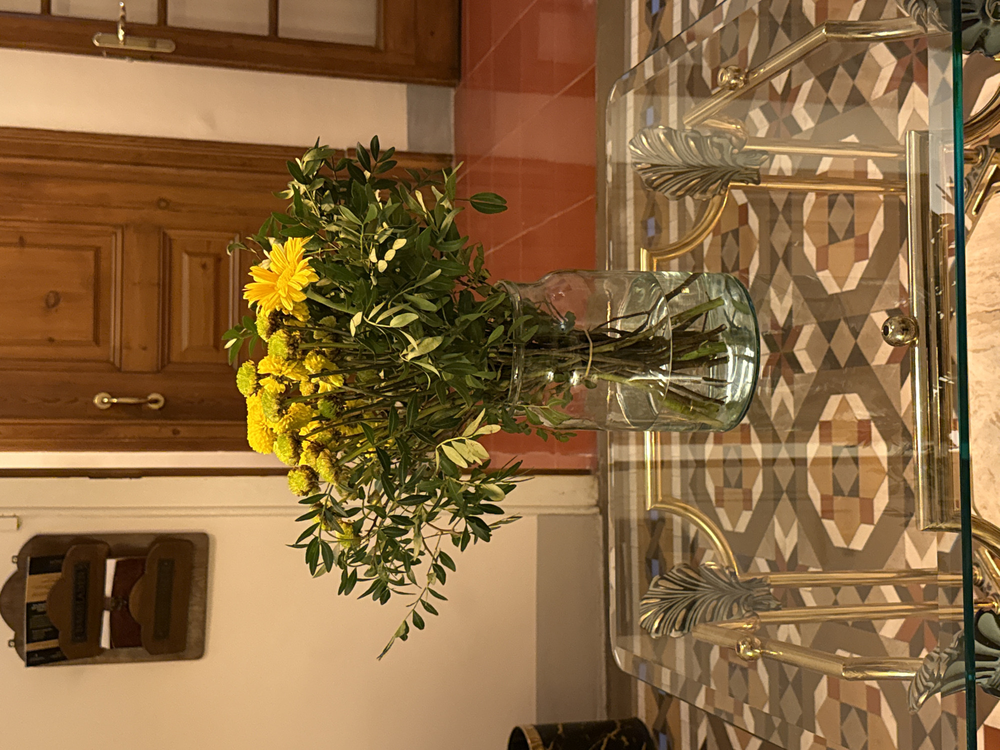 A still-life composition shows a glass table with brass trim, reflecting a patterned wall behind it, and a bouquet of yellow flowers in a glass vase on the table. The floral arrangement, dominated by yellow chrysanthemums and greenery, sits in the center of the table. The table's legs feature a brass leaf design. The background includes a dark wooden door with brass handles and a small wall-mounted shelf. The patterned wall features diamond shapes in tan, brown, and gray. The setting appears to be indoors, possibly a residence. Information about the location being in Barcelona, Spain, is not directly discernible from the image's visual content.