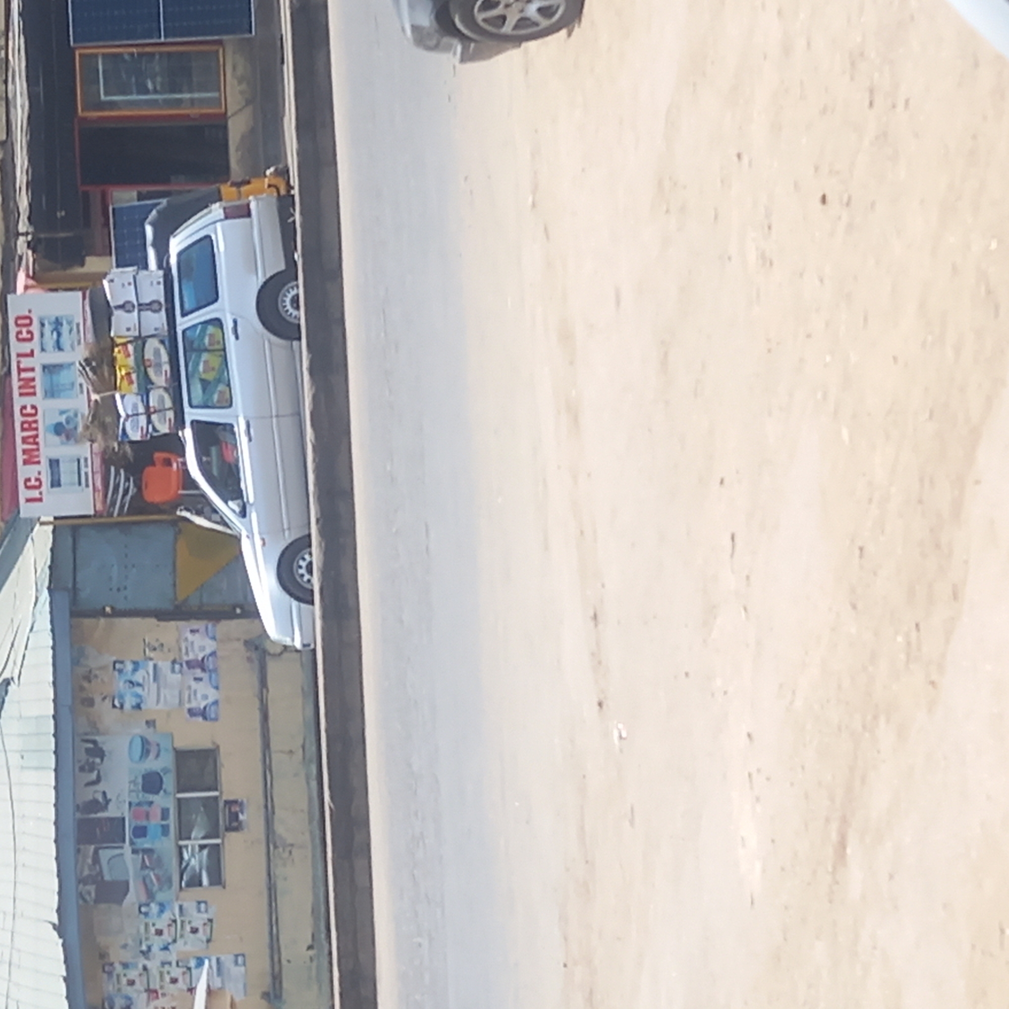 The image, rotated 90 degrees counter-clockwise, depicts an outdoor street scene, likely in Dadi Riba, Nigeria. On the right side of the frame, a white van, resembling a mini-truck or pickup, is parked on the side of a dirt or unpaved road. The van carries several boxes or packages secured on its roof rack. A bright orange canister, possibly a gas cylinder, is visible within the van's open cabin.
On the left side of the frame, a building houses a business, identifiable by a large vertical sign that prominently displays the text "I.C. MARC INT'L CO." Below this, the sign features graphics of what appear to be water dispensers or related appliances. Higher up on the building, two rectangular solar panels are mounted, suggesting the business utilizes or sells solar energy solutions. Several windows are visible on the building's facade. The lower section of the building's exterior is covered with various posters and displays showcasing an assortment of household goods or electronic items. The road surface where the van is parked consists of light-colored dirt, with a darker, narrow paved edge or curb visible closer to the building. The scene is illuminated by bright daylight, indicating a sunny time of day. The combination of commercial displays, a delivery vehicle, and solar panels paints a picture of a typical active roadside commercial area.