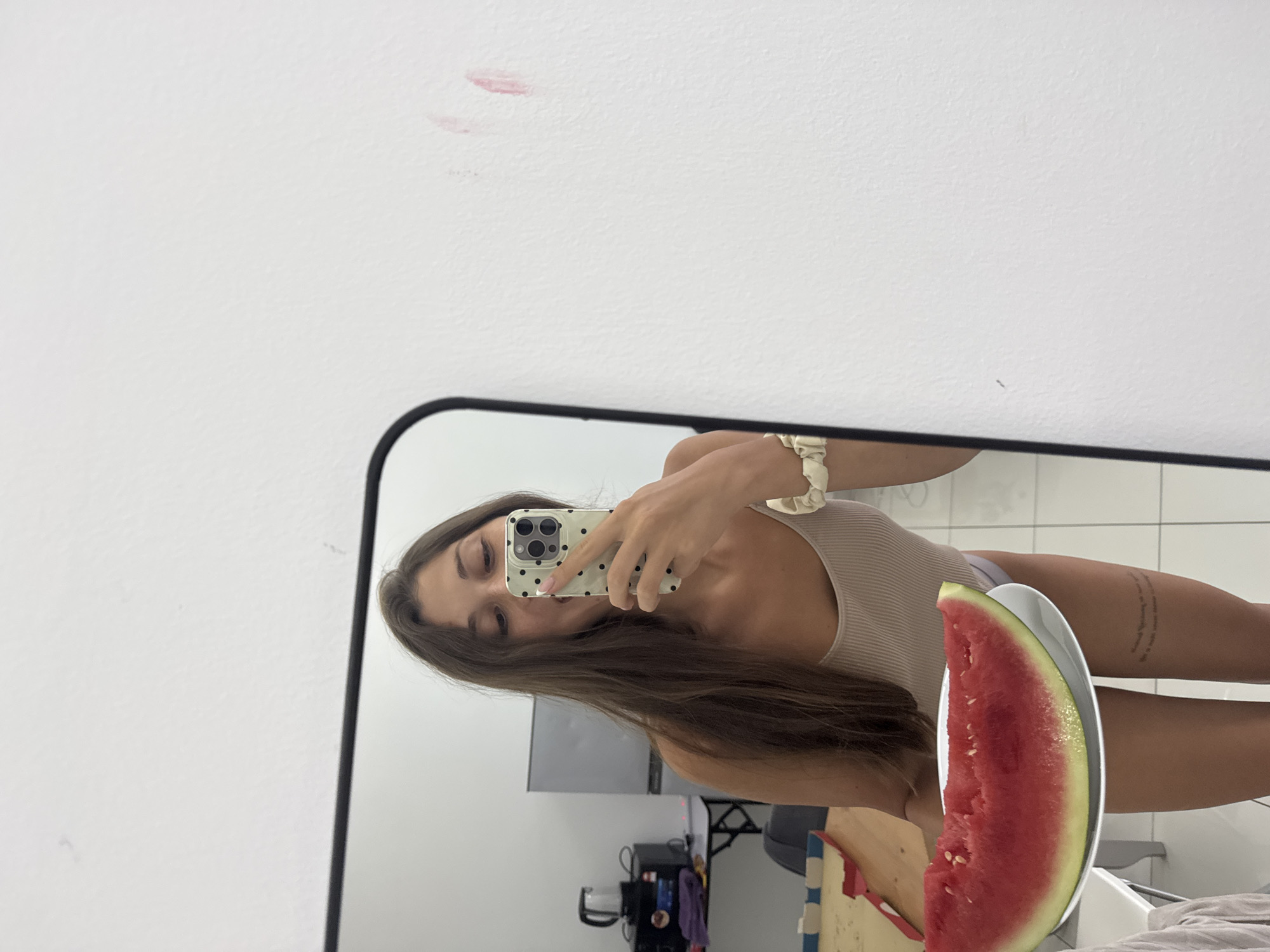 The image displays a mirror selfie of a young woman, captured with the photo rotated horizontally. The woman has long, dark brown hair and is wearing a light-colored, ribbed tank top or bodysuit and light-colored underwear. She holds a smartphone with a white case adorned with black polka dots in her right hand, using it to take the picture in the mirror. In her left hand, she holds a substantial slice of watermelon on a white plate. A cream-colored scrunchie is visible on her left wrist.
The reflection in the mirror reveals an indoor setting, consistent with a room in a residence in Kuala Lumpur, Malaysia. The background includes a white tiled floor, a dark television or monitor set on a stand, and various dark objects and cables on a surface that appears to be a cabinet or shelf. A section of what looks like a bed or draped furniture with light-colored bedding is visible on the right side of the frame.
Outside the mirror's frame, on the white wall, there are faint pink smudges, possibly lipstick marks. The woman has a multi-line text tattoo on her left thigh, though the text itself is not clearly legible. Her expression is neutral, as she is looking down at her phone screen. The lighting indicates a brightly lit indoor environment.