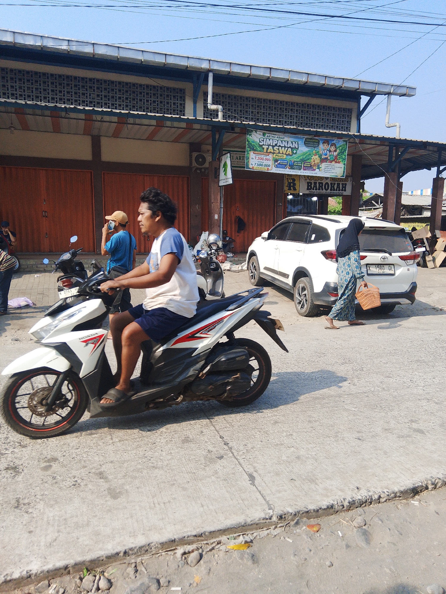 A medium shot captures a man riding a white scooter on a concrete road in front of a building with orange doors and a corrugated metal roof. He is wearing a white and blue t-shirt and dark blue shorts, with dark sandals on his feet. To his left, another man in a blue t-shirt and a beige baseball cap is talking on a cell phone. Further back, a white SUV is parked. A woman wearing a black hijab and a blue and white patterned dress is walking away from the SUV, carrying a woven basket. The building appears to be a commercial establishment, with a banner advertising "SIMPANAN TASWA" visible on its facade. The scene is set in Kalinyamatan, Indonesia, with clear blue sky and bright sunlight casting shadows on the ground.