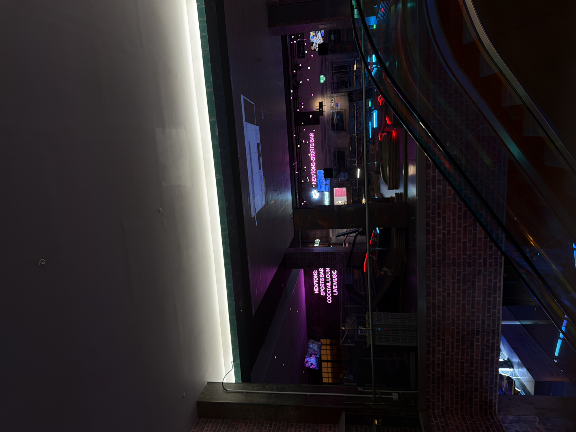 The image captures a vibrant, multi-level indoor entertainment venue, likely an establishment designed for evening activities. The scene is illuminated almost entirely by artificial light, creating a dynamic and modern atmosphere.
A dominant feature on the left is a tall, bright white illuminated wall or light strip, contrasting sharply with the darker, subdued tones of the surrounding surfaces. On the right, a sleek escalator with glass side panels and dark handrails descends alongside a textured brick wall, reflecting the colorful lights of the space.
In the mid-ground and background, the venue's purpose becomes clear through prominent neon signage. A large pink neon sign spells out "NEWTONS SPORTS BAR," with another below it reading "COCKTAIL LOUNGE" and "LIVE MUSIC." These signs indicate the primary offerings of the establishment. Numerous other colorful lights, including blue, red, and additional pink accents, are visible throughout the space, suggesting screens, decorative lighting, or other neon displays. Some small, round pink lights are strung across an area, adding to the festive ambiance. A faint projection, possibly of a gaming console, is visible on a flat surface in the background.
No people are present in the image, but the arrangement of signs and lights suggests a lively social hub. The lighting conditions imply it is either nighttime or an interior designed to simulate a nighttime experience. The architecture blends modern elements like glass and bright light strips with traditional materials such as brick.