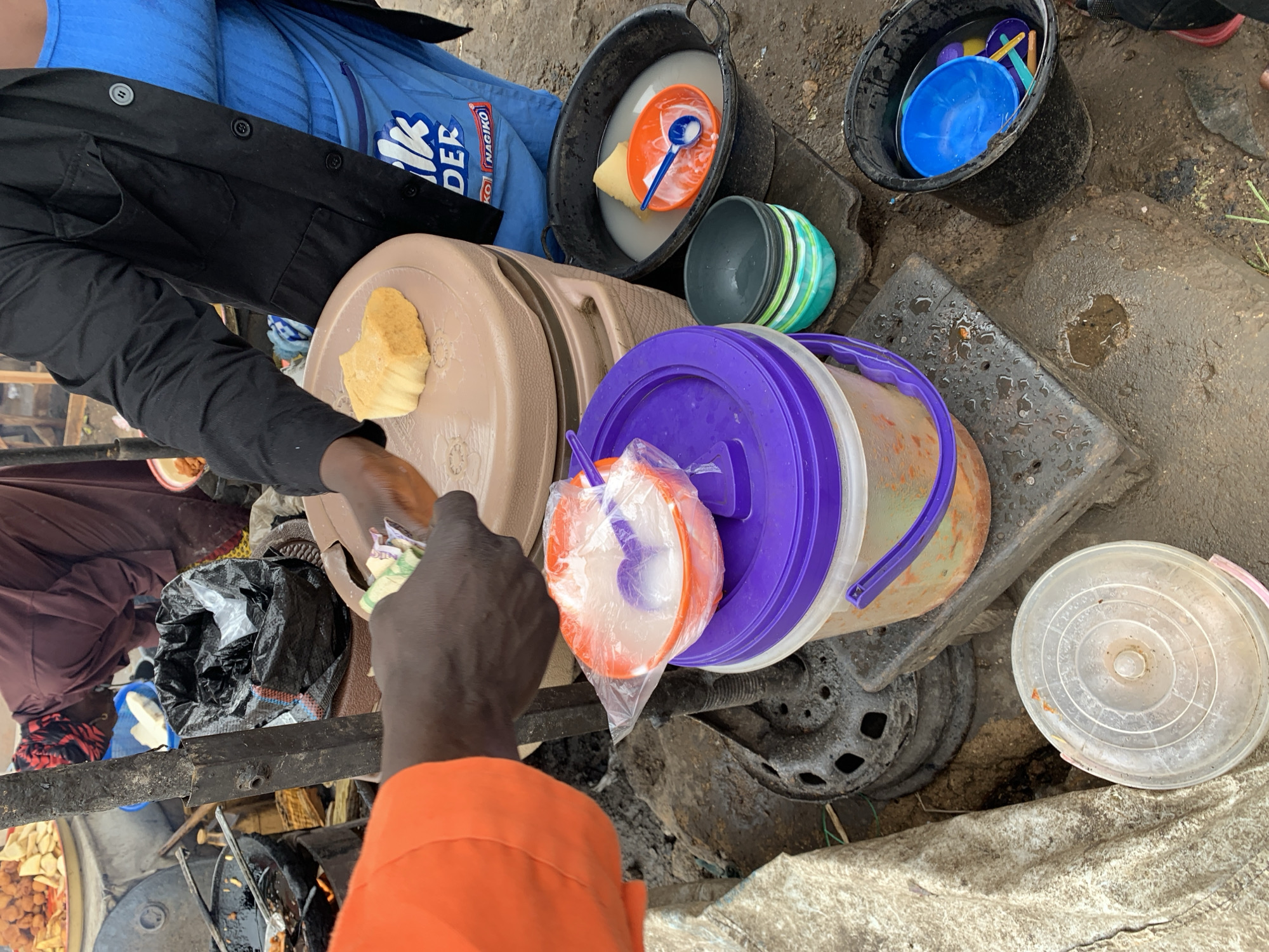 The image depicts a street vendor scene, likely in Rafin Par, Nigeria, based on the cultural context of food preparation and items. An individual with dark skin is exchanging currency for food, possibly a fried dough variant, which appears to be served from a large beige container. The vendor is positioned beside an array of buckets and containers, including one bucket with blue utensils, and another with a white liquid and a spoon, indicative of washing or food preparation materials. The environment consists of rough, potentially unpaved ground. A visible "NAGIKO" logo on clothing suggests a connection to a local brand. There's evidence of cooking equipment and fried food, potentially akara, further cementing the street food vending scene. The scene overall presents a basic informal food commerce environment.
