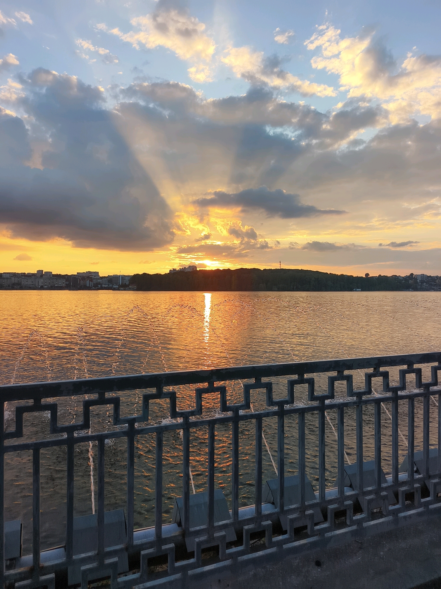 The image captures a sunset over the Ternopil Reservoir in Ternopil, Ukraine. The sun is low on the horizon, casting a warm golden glow across the water and illuminating the clouds with orange and yellow hues. Rays of sunlight pierce through the scattered clouds, creating a dramatic effect. In the distance, the shoreline is lined with trees and buildings, suggesting an urban or suburban setting. In the foreground, a metal railing with a geometric pattern separates the viewer from the water. The water itself is rippled, reflecting the colors of the sky and creating a shimmering surface. Several thin, vertical streams of water, possibly from a fountain, are visible in the middle ground, breaking the surface of the water with splashes. The scene is serene and picturesque, typical of a sunset over a body of water.
