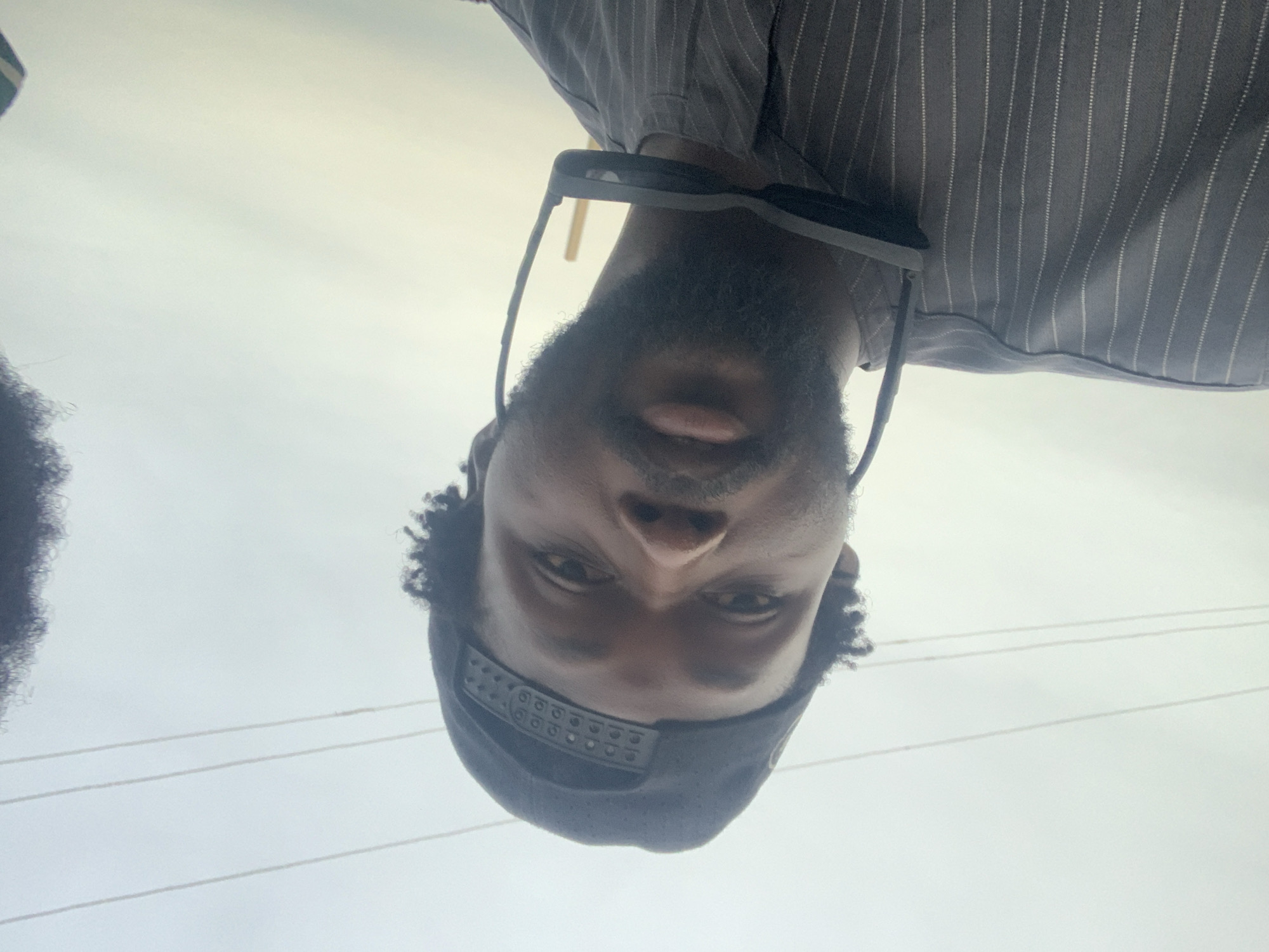 The image is a close-up, low-angle selfie of a Black man wearing a black baseball cap and a grey pinstripe shirt. Sunglasses are hanging from the collar of his shirt. He has a beard and his hair is curly. The background is a pale, overcast sky with two horizontal power lines visible. The scene is outdoors, suggesting it was taken in Takum, Nigeria.