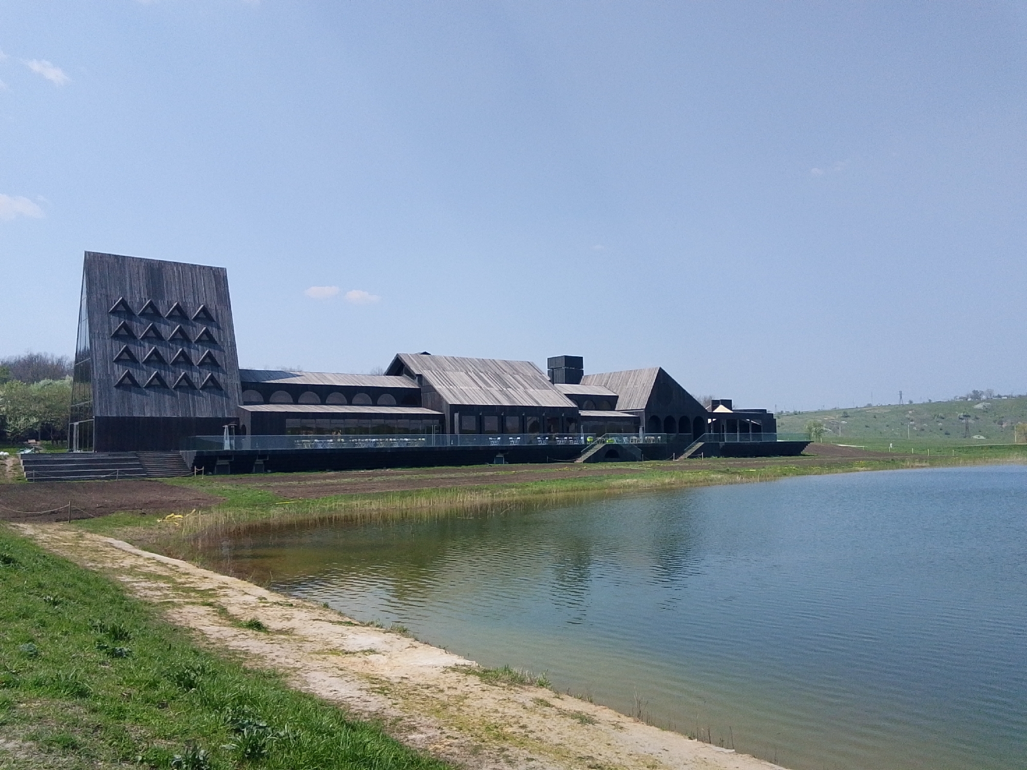 The image shows a large, modern building made of dark wood situated next to a body of water, likely a lake or pond. The building has a unique architectural style, featuring a tall tower-like structure with triangular openings and a long, low section with a gabled roof. The building appears to have a deck or patio extending over the water.
In the foreground, there is a grassy shoreline with a sandy bank. The water is relatively calm with visible ripples. In the background, there is a grassy hillside. The sky is clear and blue with a few small clouds.
The image provides no evidence of people or interactions taking place within the scene.
The location is identified as Каменка, Russia.