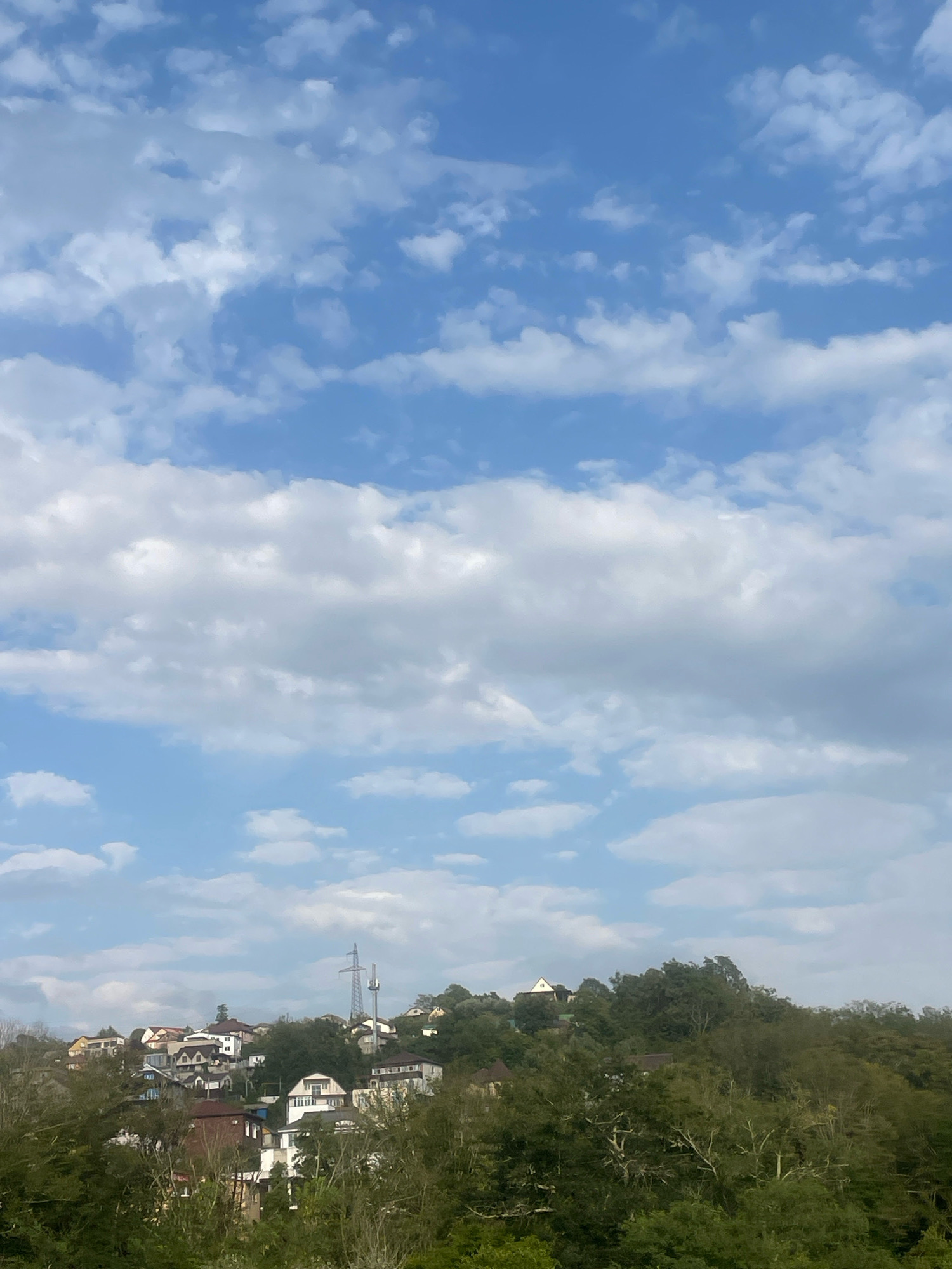 The image depicts a view of a hillside residential area in Sochi, Russia, with a sky filled with white clouds. The foreground is dominated by lush green trees, partially obscuring the view of the settlement. Several houses with varied architectural styles and roof colors are scattered across the hillside, interspersed with dense foliage. A prominent power pylon with two antennas stands tall on the ridge, indicating the presence of infrastructure. The sky above is a clear blue, with scattered cumulus clouds contributing to a dynamic sky. The overall scene suggests a tranquil, possibly suburban or semi-rural environment within Sochi.