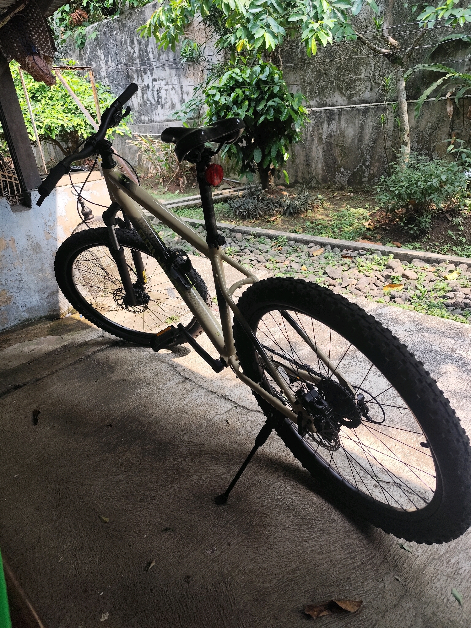 A tan-colored mountain bike with black tires and a black seat is parked on a concrete surface. The bike is leaning on its kickstand. The background shows a concrete wall, lush green foliage, and some rocks. The scene appears to be in an outdoor, possibly residential, area. The lighting suggests it is daytime, with sunlight casting shadows on the ground. The overall environment suggests a tropical or subtropical climate. This scene is consistent with an outdoor space in Bogor, Indonesia, characterized by its humid climate and abundant vegetation.