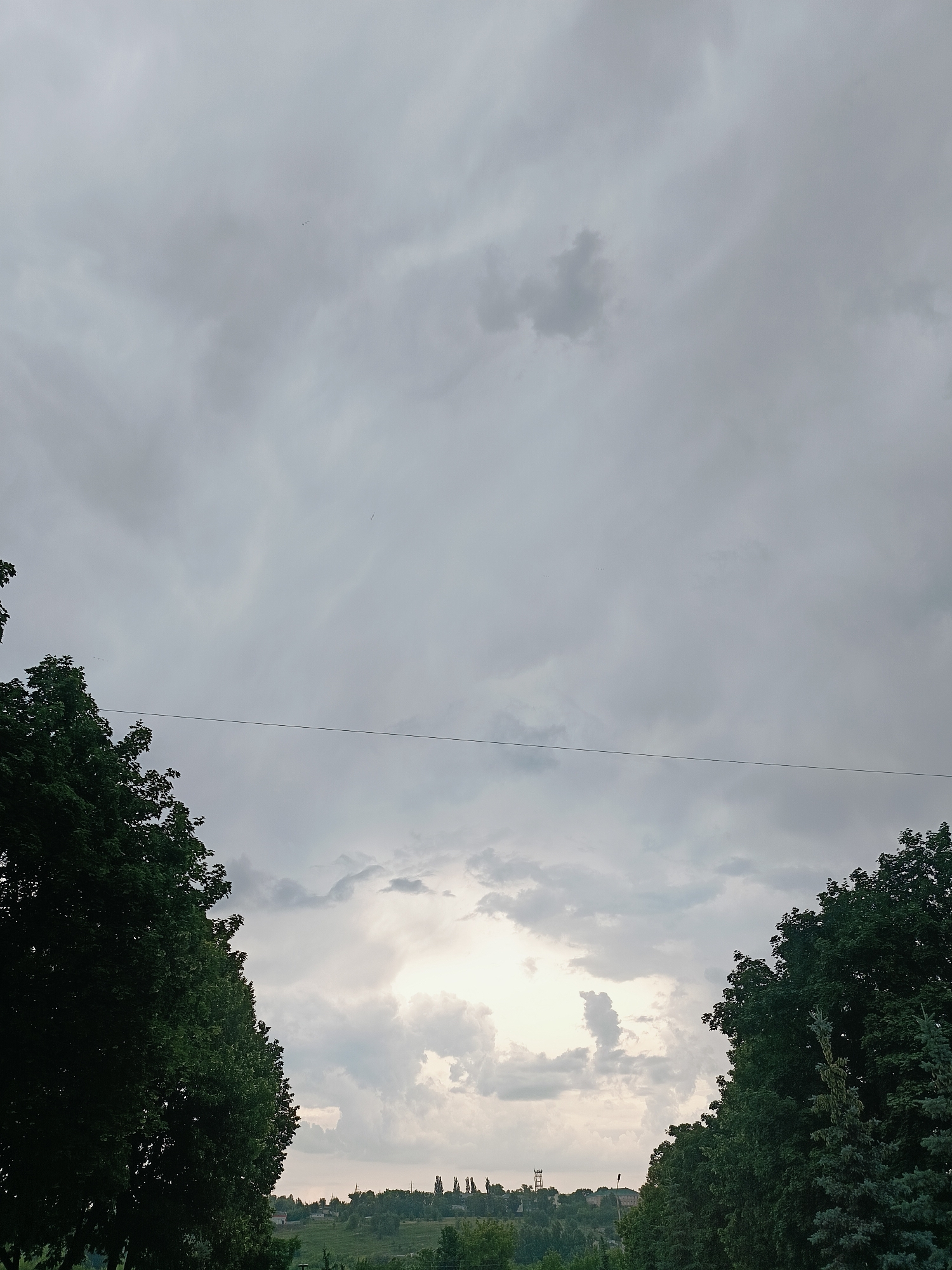 The image depicts a wide shot of a cloudy sky over a landscape of rolling hills and trees. In the foreground, on the left and right sides of the frame, are large, dark green trees. A thin, dark horizontal line, likely a wire, stretches across the middle of the image. The sky is overcast with various shades of gray and white clouds, with a patch of brighter light visible in the distance towards the center. In the background, a hazy outline of buildings and trees suggests a settlement or town. The scene appears to be a natural landscape in or near Gubkin, Russia, on a cloudy day. There are no people, animals, or specific actions visible in the image.