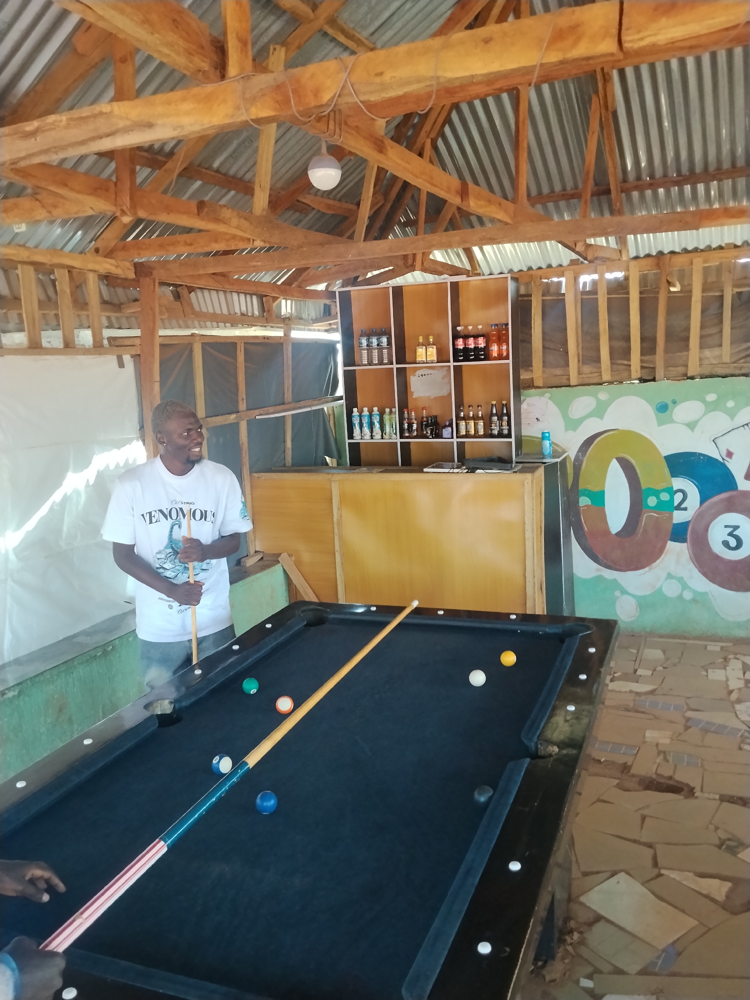 The image depicts an indoor recreational area with a pool table in the foreground. A young man with bleached hair, wearing a white t-shirt and jeans, stands behind the pool table, holding a cue stick. He is looking to his right and smiling. Several pool balls are scattered on the black felt surface of the table, including a green ball, a white ball, a yellow ball, and a blue ball. A long cue stick rests on the table, extending from the foreground towards the man.
In the background, a wooden shelf unit is filled with various bottles, likely beverages. To the right of the shelf unit, a colorful mural is painted on the wall, featuring large numbers such as "0", "2", and "3", and stylized pool ball graphics. The ceiling is constructed with wooden beams and corrugated metal sheeting. The overall environment suggests a casual, outdoor-style establishment. The location is identified as Durimi, Nigeria.