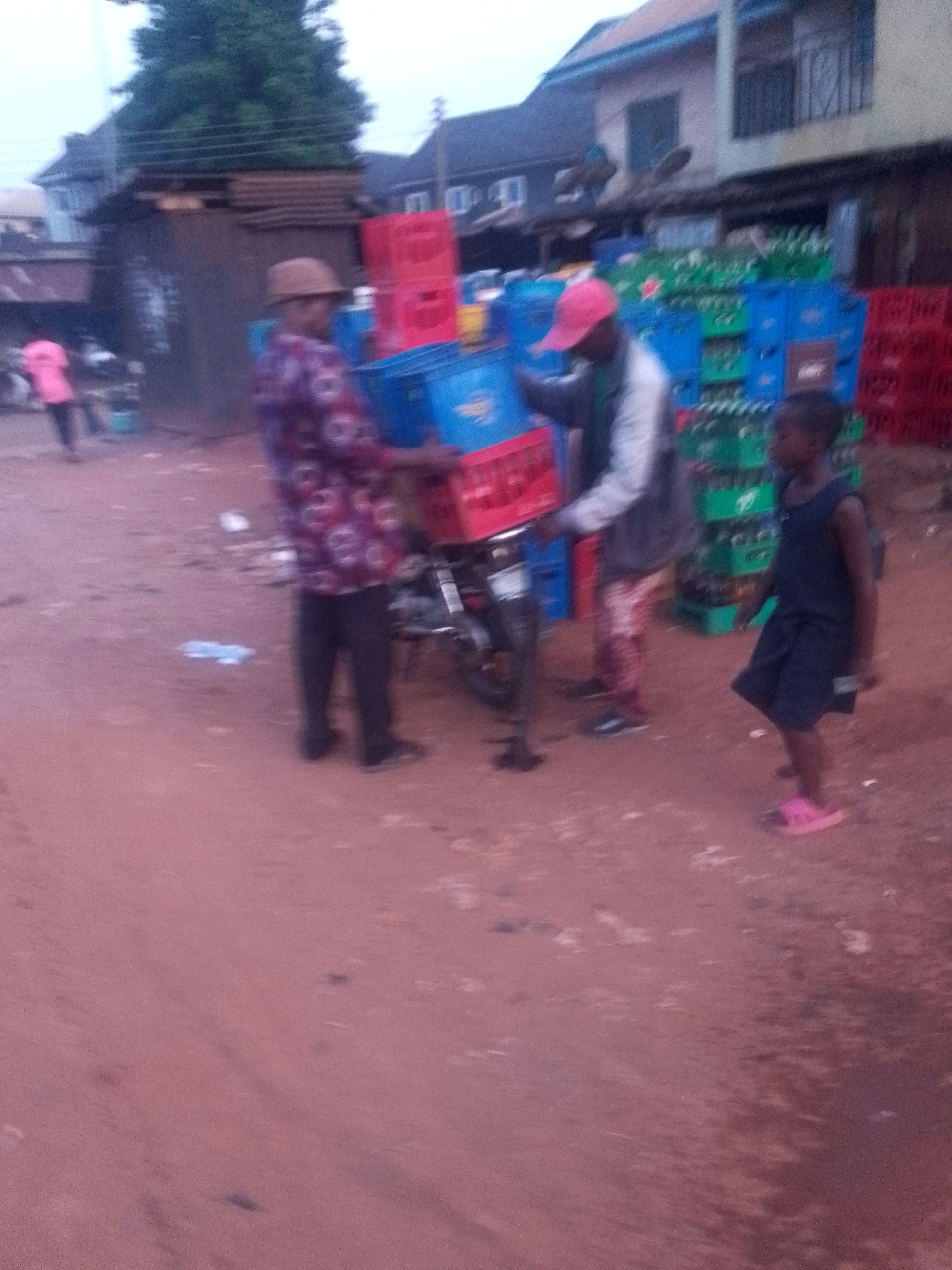 In an outdoor scene in Unknown, Unknown, two men are loading stacks of colorful plastic crates onto a motorcycle. The crates are predominantly red, blue, and green. One man wears a brown hat and a patterned purple and red shirt, while the other sports a red cap and a light-colored jacket. A child in a dark outfit and pink sandals stands nearby. The ground is reddish dirt, and buildings are visible in the background.