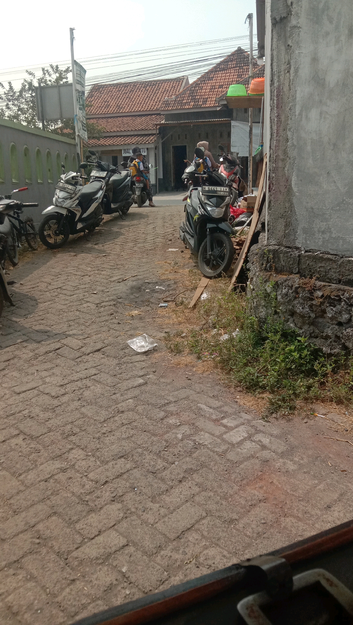 The image depicts a street scene in Karang Anyar, Indonesia, characterized by a cobbled pathway leading towards a row of buildings with terracotta roofs. Several parked motorcycles are visible, some with riders. In the foreground, a concrete wall and some overgrown vegetation are present. The scene appears to be a busy area, likely a commercial or residential street. The overall impression is one of a typical day in a moderately developed urban environment.