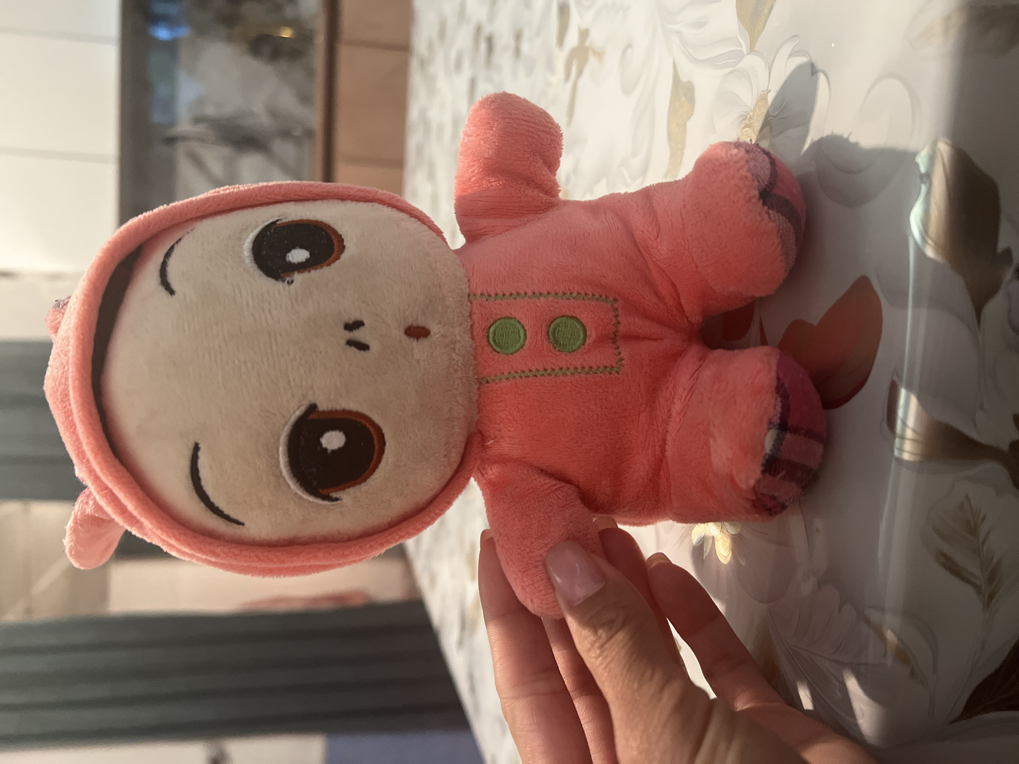 A close-up, eye-level shot shows a plush toy with a pink hooded onesie sitting on a table in Chong-Sary-Oy, Kyrgyzstan. The toy has large brown eyes, a small nose, and a surprised expression with its mouth open. It has two green buttons on its chest, and its feet have a plaid pattern of pink and dark purple stripes. A person's hand, with neatly manicured nails, is gently holding the toy's arm. The table is covered with a patterned tablecloth, and the background is blurred, showing hints of a domestic interior with cabinets and what appears to be a reflection of lights and possibly a television screen.