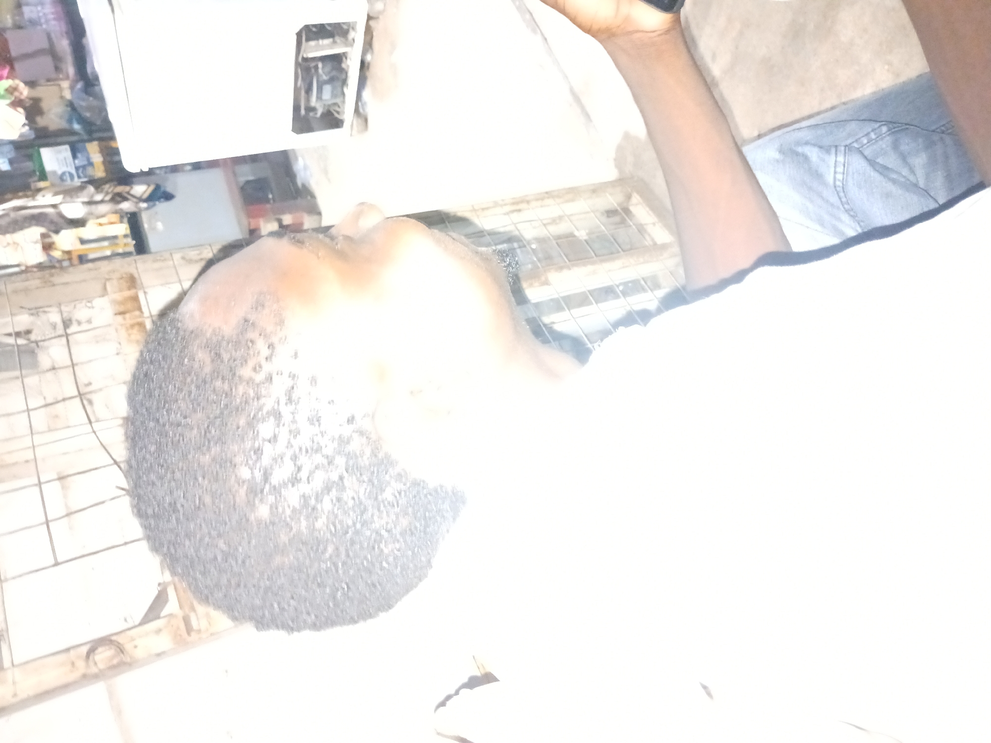 The image depicts a close-up of a person's head and shoulder, with the background showing what appears to be a dimly lit interior space. The person has short, dark, curly hair and is wearing a white shirt and blue jeans. Their right arm is visible, with a dark-colored object, possibly a watch or bracelet, on their wrist. In the background, there is a white appliance, possibly a freezer, with its back panel open, revealing internal components. There is also some shelving with various items, and a caged structure. The overall lighting is low, with some areas appearing overexposed. The image quality is poor, with significant digital noise. There is no visible indication of the city of Jalingo, Nigeria.