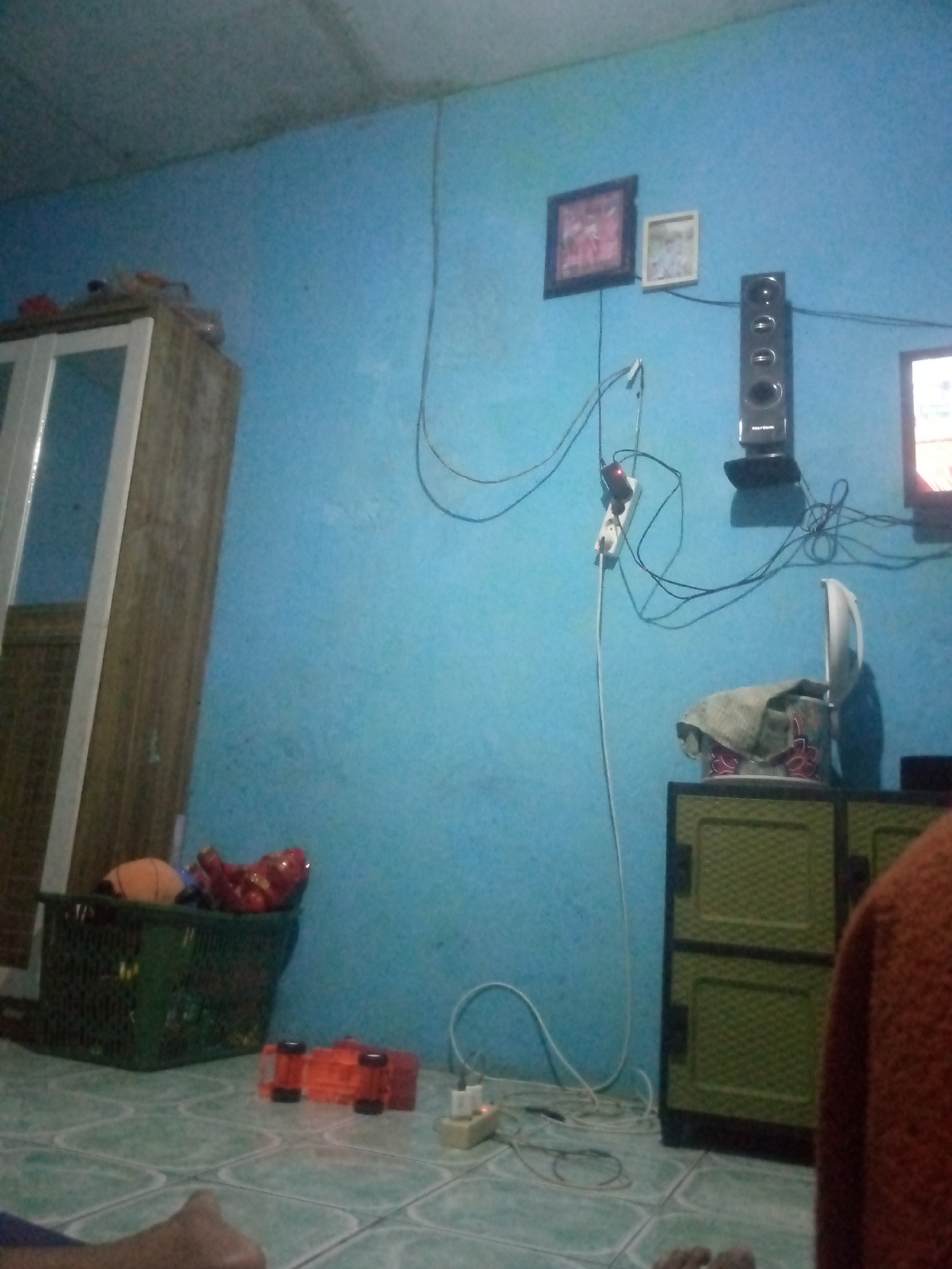 The image depicts an indoor scene, likely a room in a residential dwelling in Giriasih, Indonesia. The walls are painted a vibrant blue, and the floor is tiled with a greenish-blue pattern. In the background, a tall, dark speaker stands on a black base, mounted on the wall. To the left of the speaker, two framed pictures hang on the wall, one with a dark frame and the other with a lighter frame. Several electrical cords are visible, including one running from a power strip mounted on the wall, which is connected to the speaker.
In the foreground, a dark green plastic basket is filled with various items, including what appears to be a basketball and some other indistinct objects. To the right of the basket, an orange toy truck lies on its side on the floor. Further to the right, a green plastic cabinet with two doors is partially visible. On top of the cabinet, a white rice cooker with a patterned lid is covered with a light-colored cloth. A person's foot and lower leg are visible in the bottom left corner of the frame, and a portion of another person's torso wearing an orange garment is visible in the bottom right. The overall impression is of a cluttered living space.