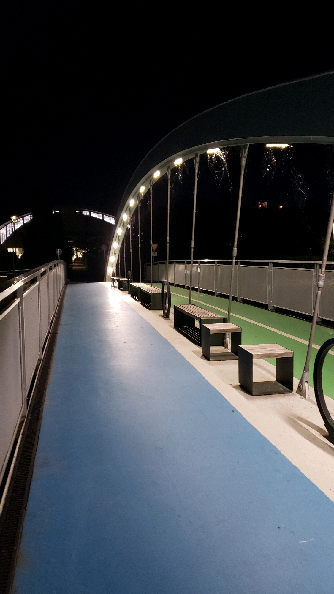 The image depicts a pedestrian and cycling bridge at night. The bridge has a blue pedestrian lane and a green cycling lane separated by a white line. Several modern, dark metal benches with wooden seats are positioned along the cycling lane. The bridge is illuminated by a series of lights mounted on an arching structure above. In the background, distant city lights and structures are visible. The overall scene is clean and well-maintained, suggesting a public space designed for recreation. There are no people visible in the image. The style of the bridge and its lighting suggest it may be a contemporary architectural feature.