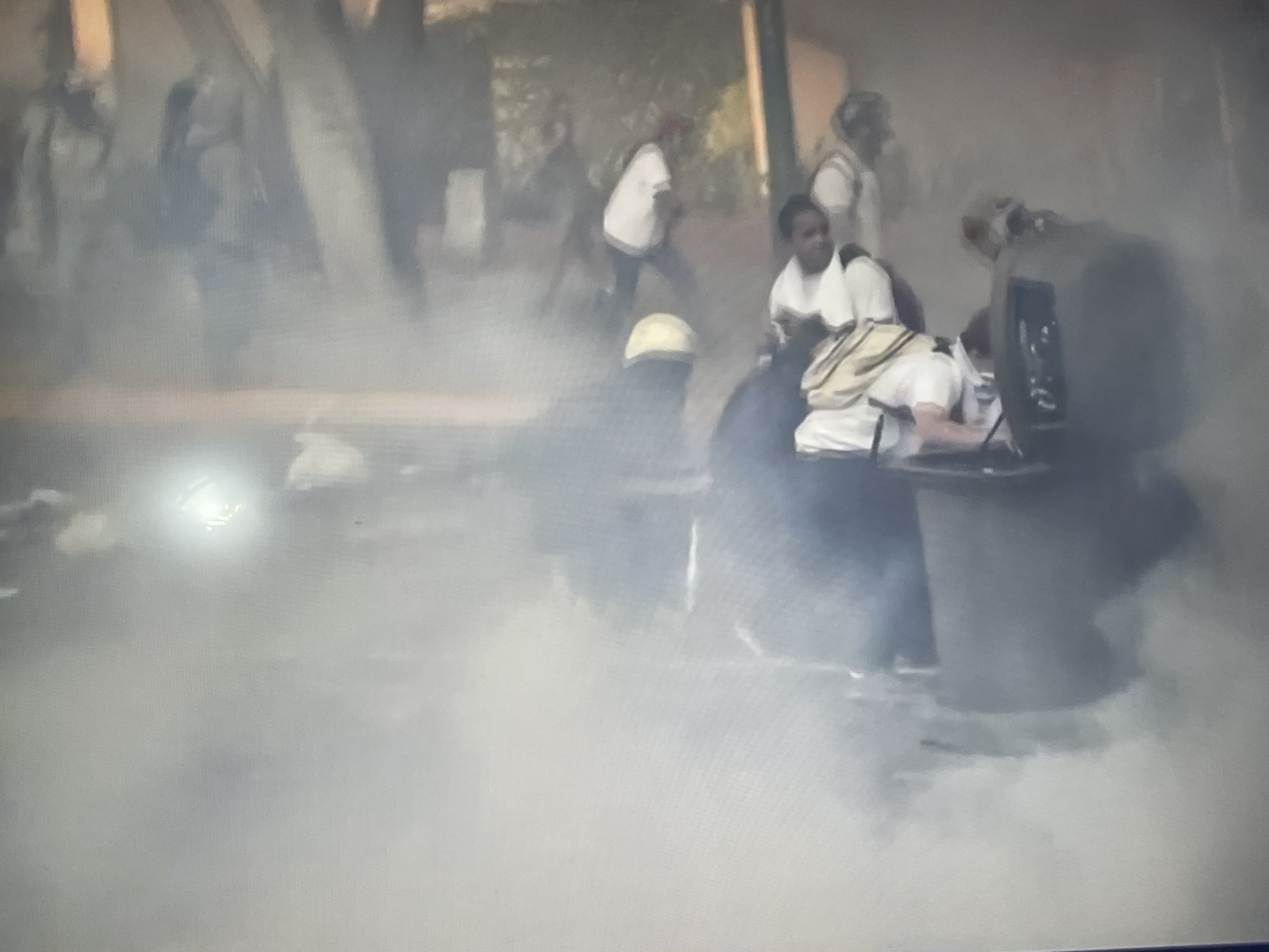 The image depicts a scene in what appears to be an urban area obscured by smoke or a haze. In the foreground, there is a dark, indistinct object with figures nearby; one individual is seated within it, and another stands close by. Several people, all wearing light-colored shirts, are visible in the background, moving away from the main focus of the image. The location is denoted as "Unknown, Unknown," suggesting an unidentified urban setting.