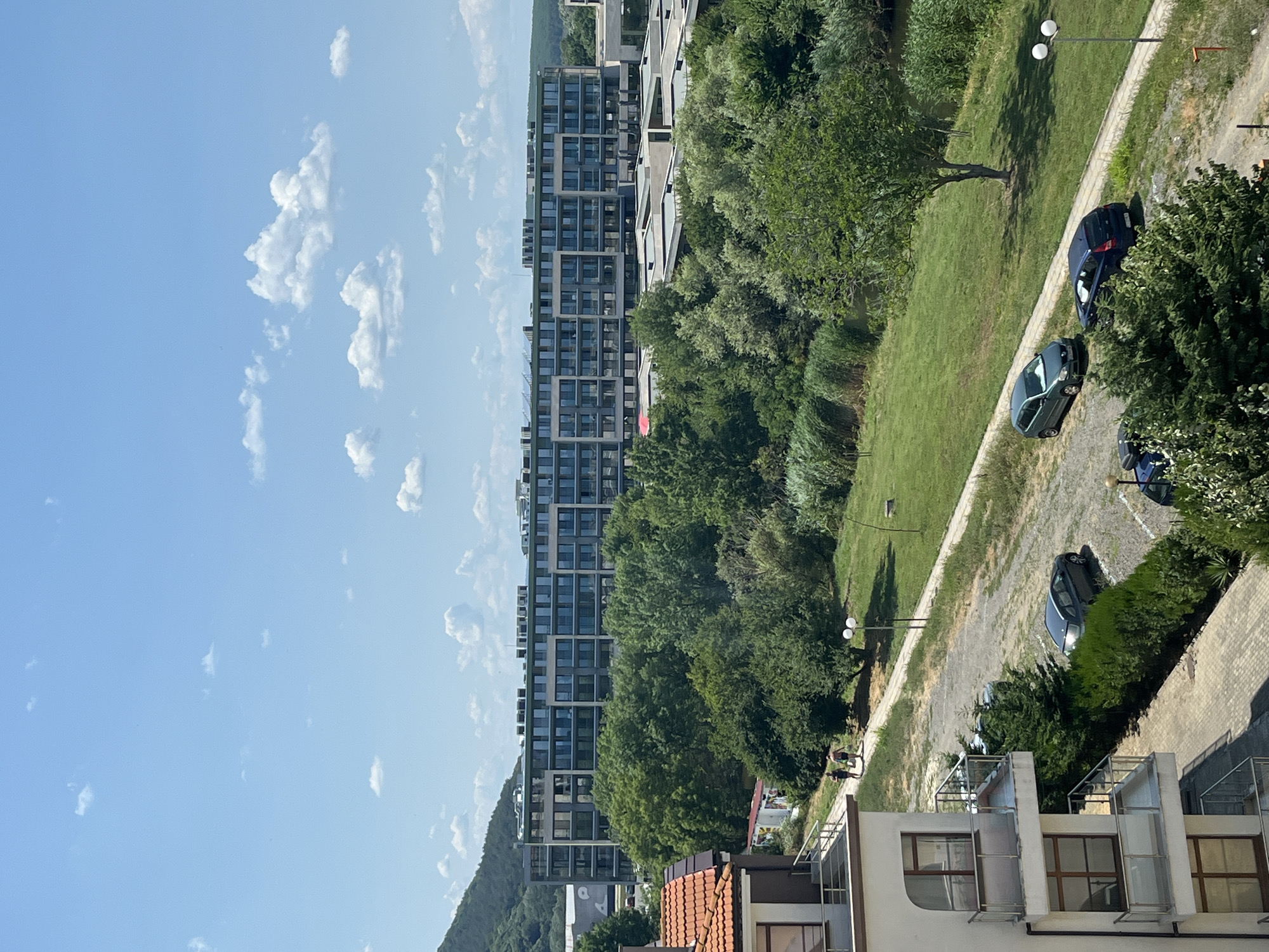 The image shows an outdoor scene with a modern apartment building and lush greenery. The building is tall and rectangular, with many windows and balconies. Trees and grass surround the building, and a paved path runs alongside it. Several cars are parked on the path. The sky is blue with scattered white clouds, suggesting a sunny day. The lighting indicates it is daytime, likely late morning or early afternoon. The overall atmosphere is serene and peaceful, with a touch of urban development blending into nature. The location appears to be a residential area with a focus on modern architecture and abundant natural elements.